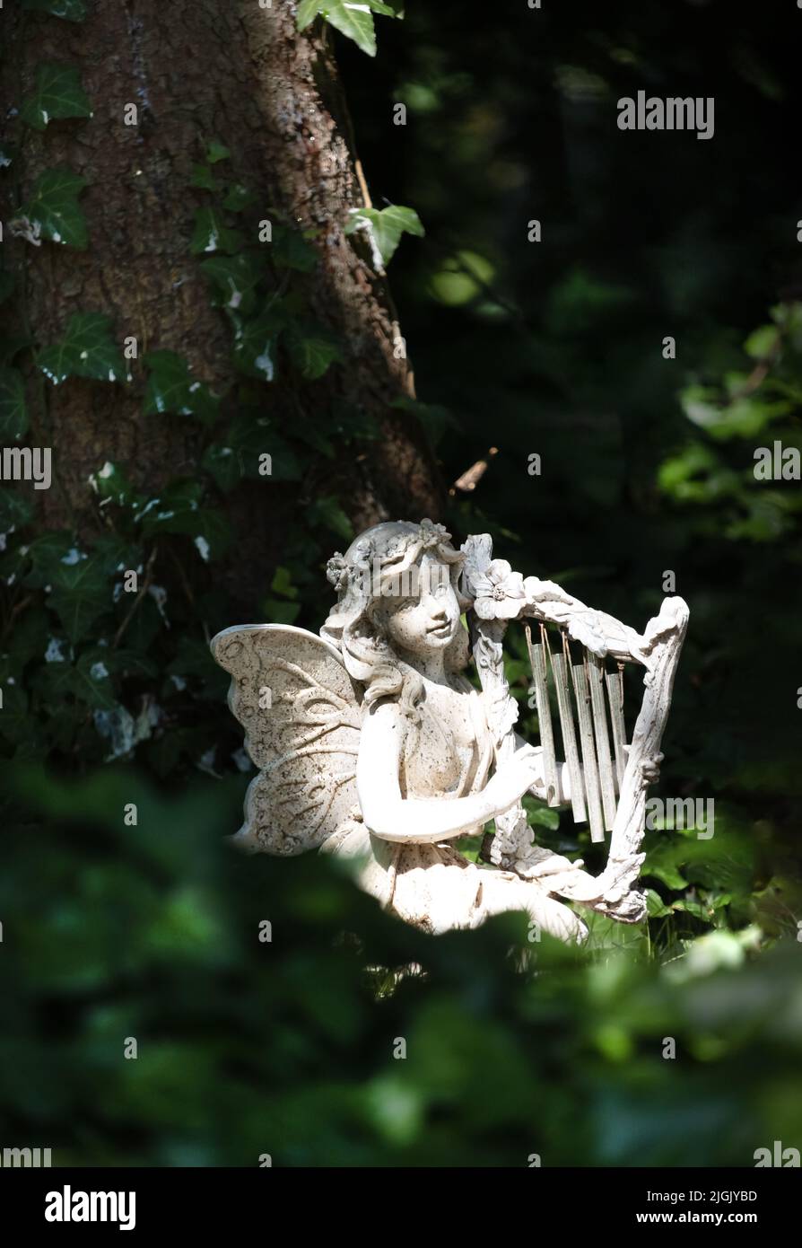 A garden statue of a fairy with a harp sitting in the woods among vines and next to a tree in spring or summer, Lancaster, Pennsylvania Stock Photo