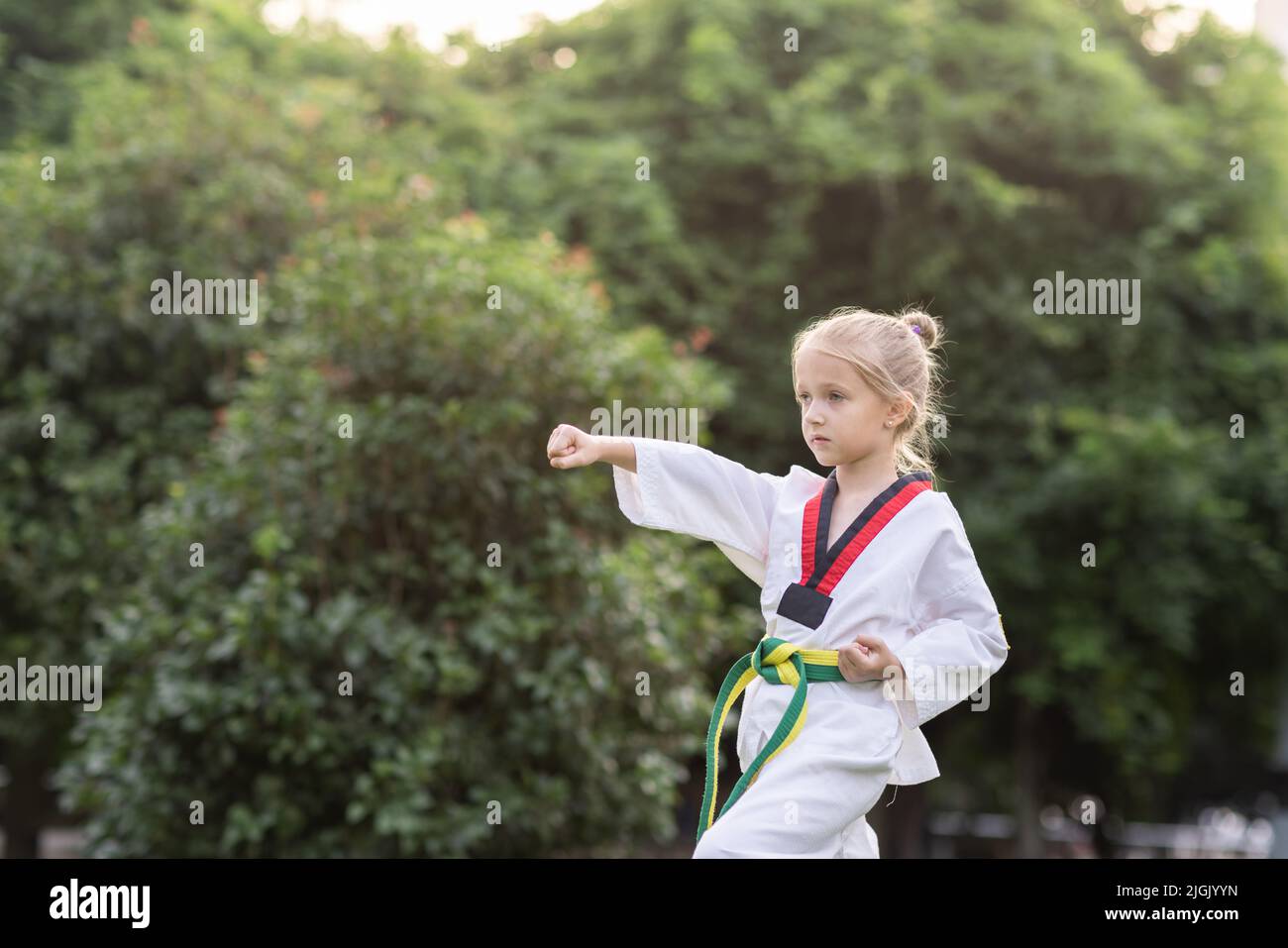 Caucasian little girl seven years old in kimono with yellow green belt exercising Taekwondo at summer park alone during coronavirus covid-19 lockdown Stock Photo