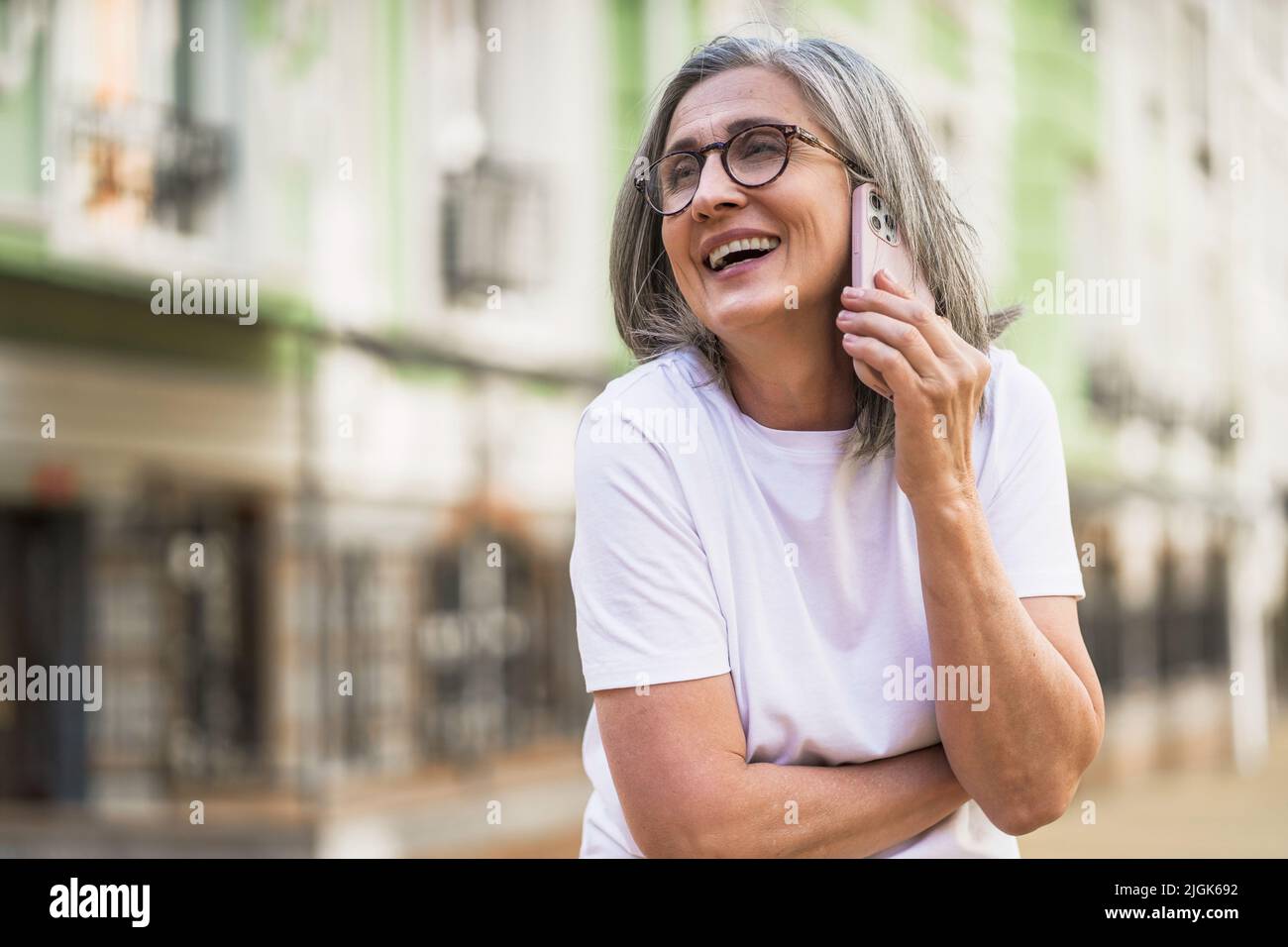 Charming mature business woman with grey hair talking on the phone holding smartphone standing outdoors of the streets of old urban city wearing white t-shirt. Silver hair woman outdoors.  Stock Photo