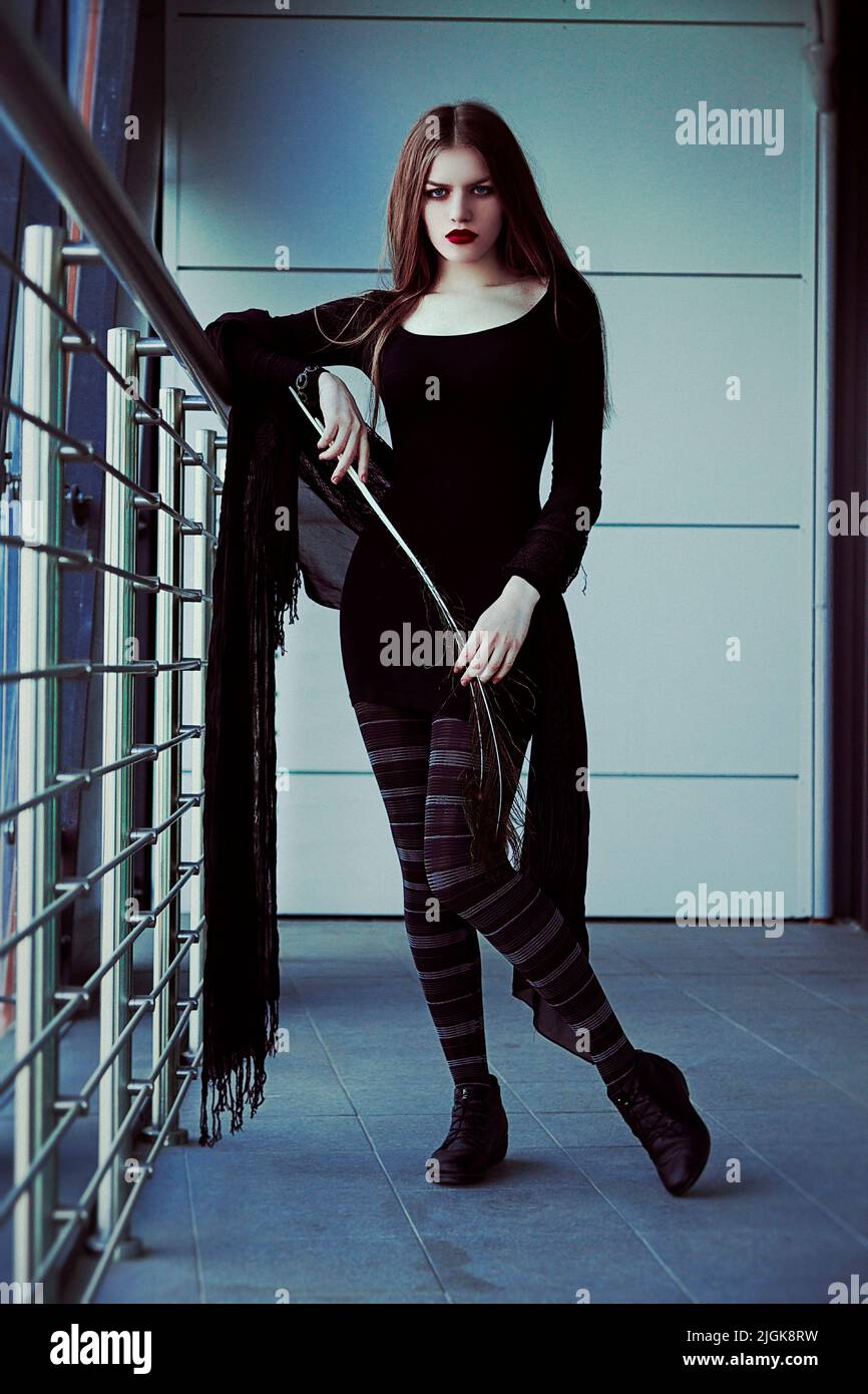 Pretty expressive brunette wearing only black clothes, holding a peacock  feather in her hand. She stands near a balcony railing; leans on it with arm Stock Photo