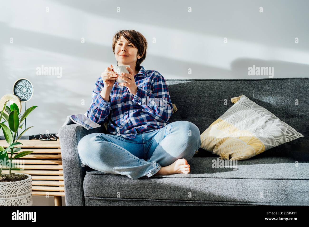 Deep in thoughts, dreaming woman drinking coffee sitting on sofa in sunlight beams after reading a book at home in cozy modern interior with green pla Stock Photo