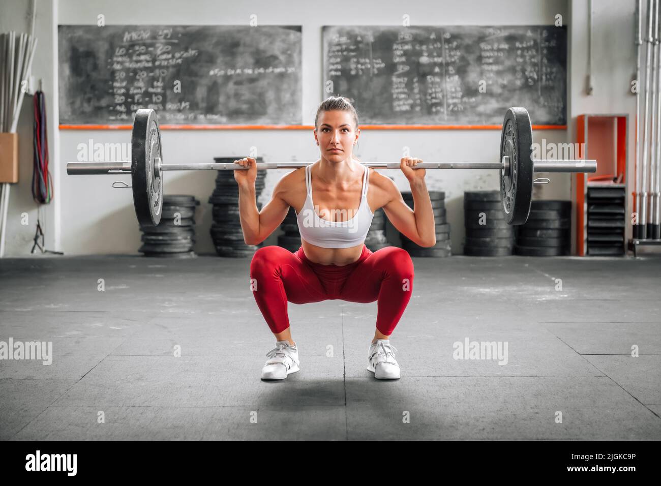 Fit female weightlifter in leggings and bra doing barbell back squat during functional workout in spacious gym in daytime Stock Photo