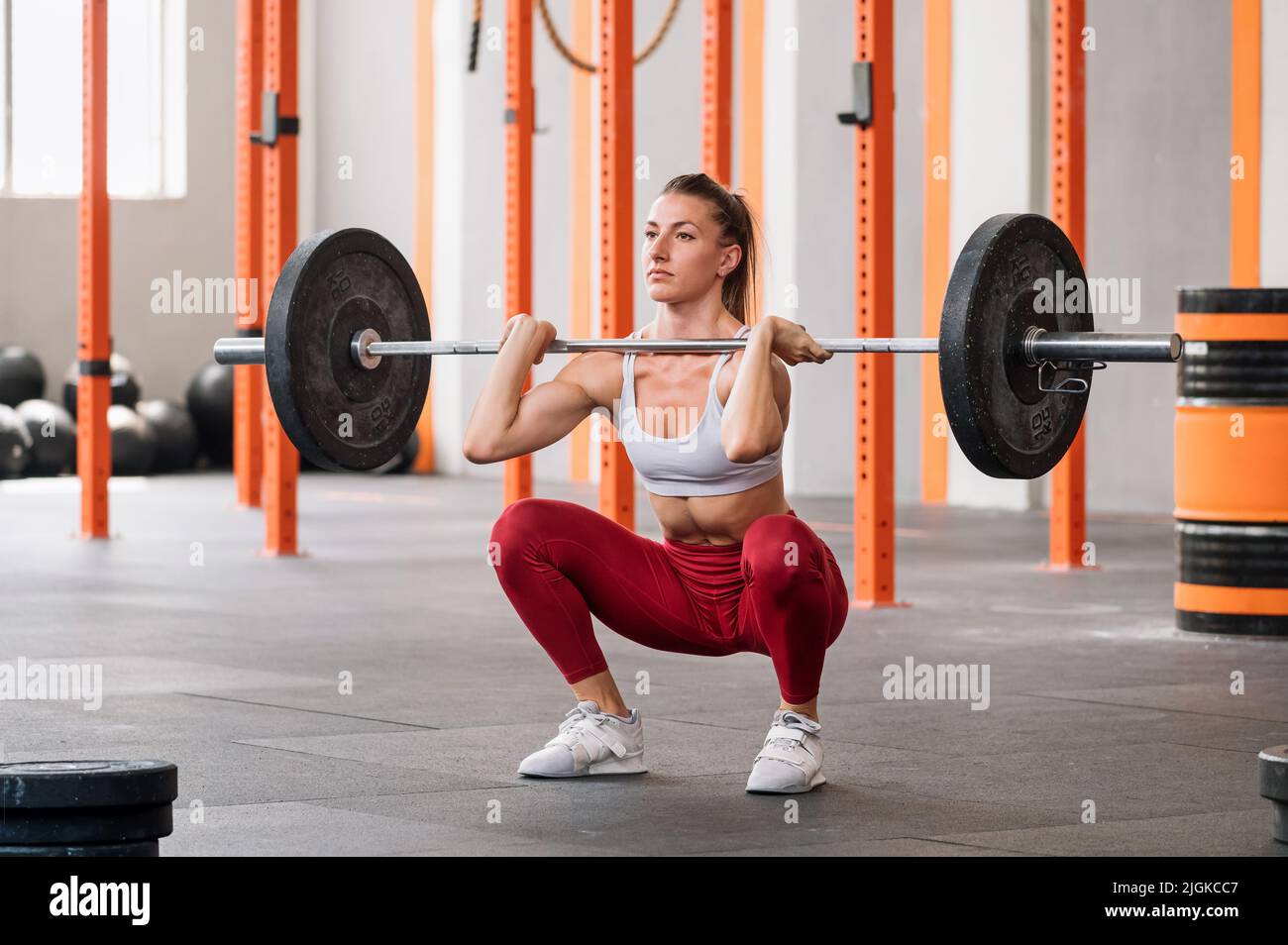 Determined sportswoman in activewear with ponytail looking away and doing front squat exercise with barbell during weightlifting workout in spacious g Stock Photo
