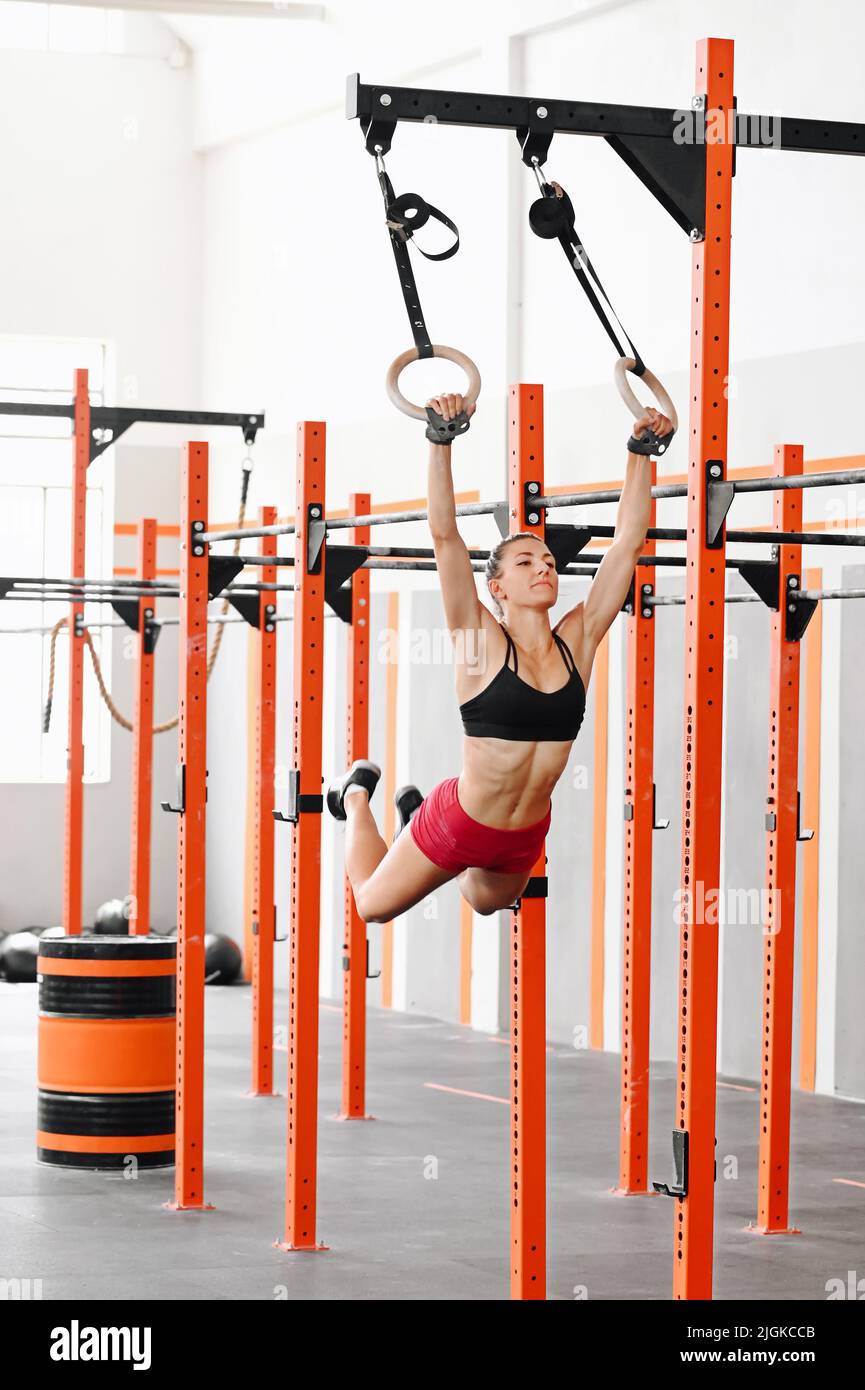 Strong sportswoman in bra and shorts swinging on gymnastic rings while doing muscle up exercise during calisthenic workout in daytime in gym Stock Photo