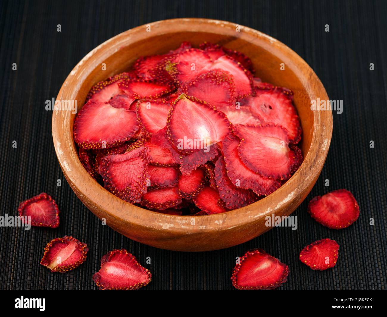 Dried strawberry slices in a wooden bowl on a black bamboo napkin. Some strawberry slices lying near Stock Photo