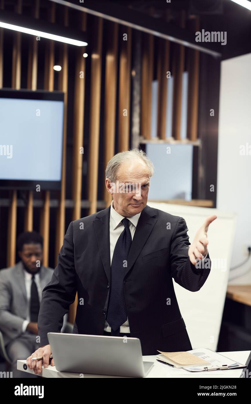 Content charismatic senior businessman in black suit standing at speech table gesturing hand at forum participant while asking his opinion Stock Photo