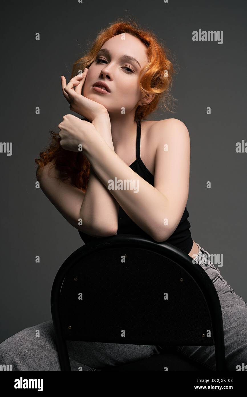 Pretty young redhead lady sitting on chair leaning at hand and bored look at camera Stock Photo
