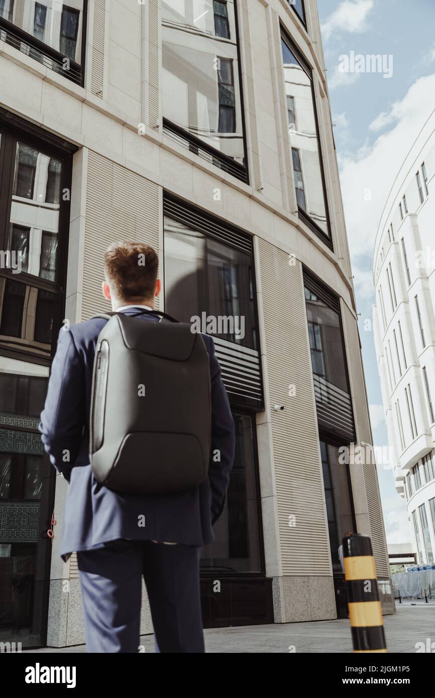 Young clerk in a suit with a backpack stands in front of a corporate building and looks up, back