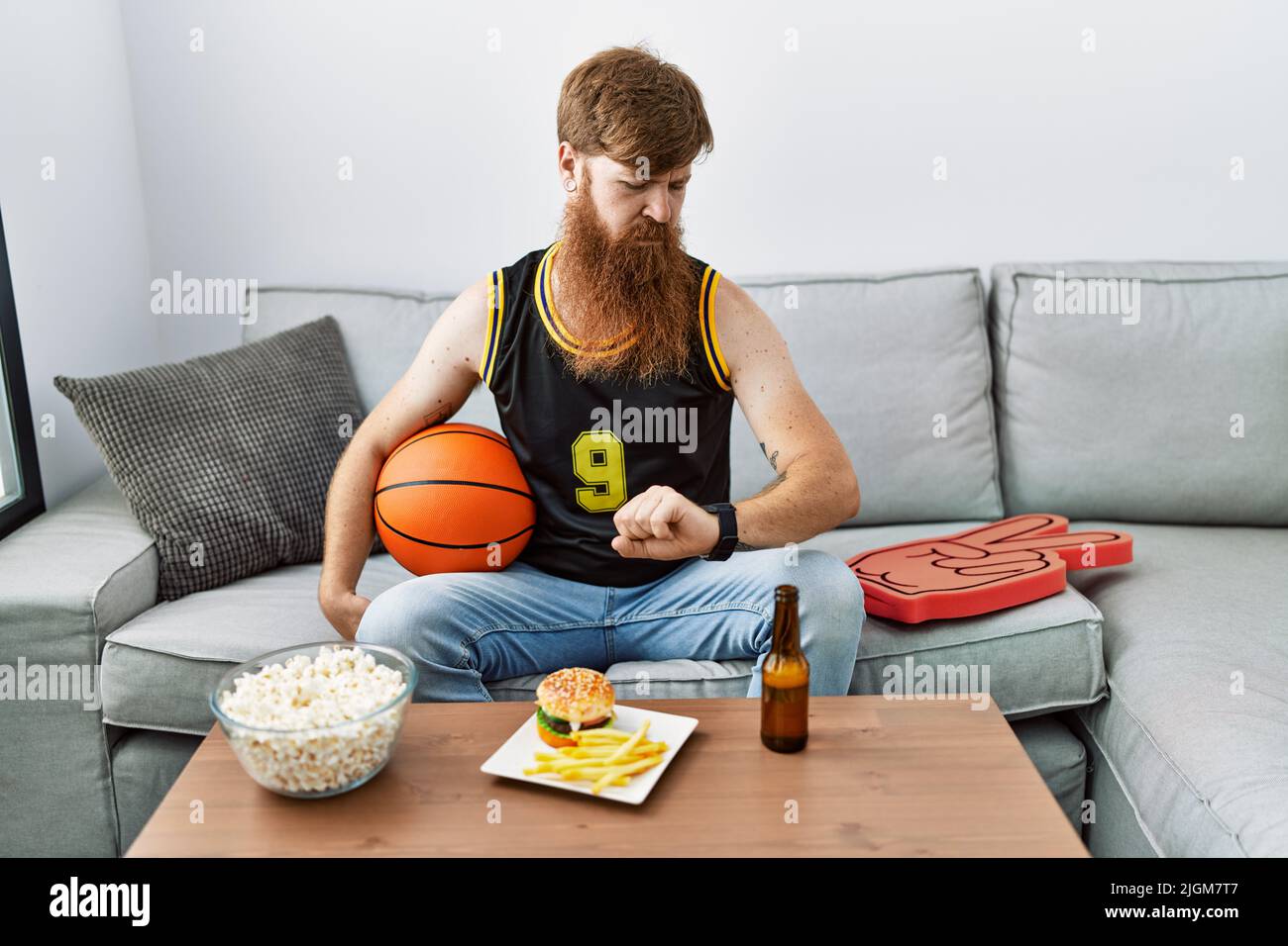 Caucasian man with long beard holding basketball ball cheering tv game checking the time on wrist watch, relaxed and confident Stock Photo