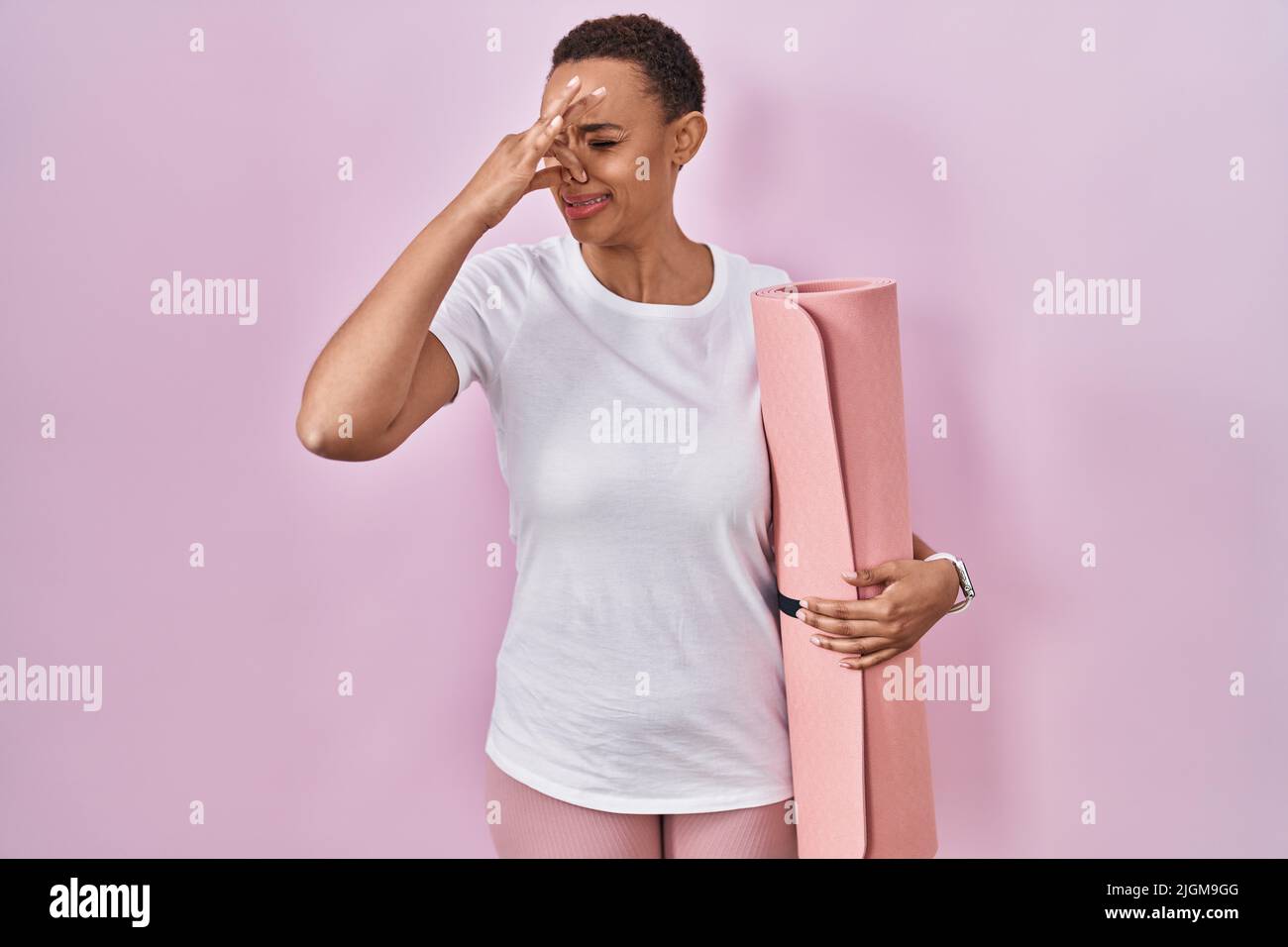 Beautiful african american woman holding yoga mat smelling something stinky and disgusting