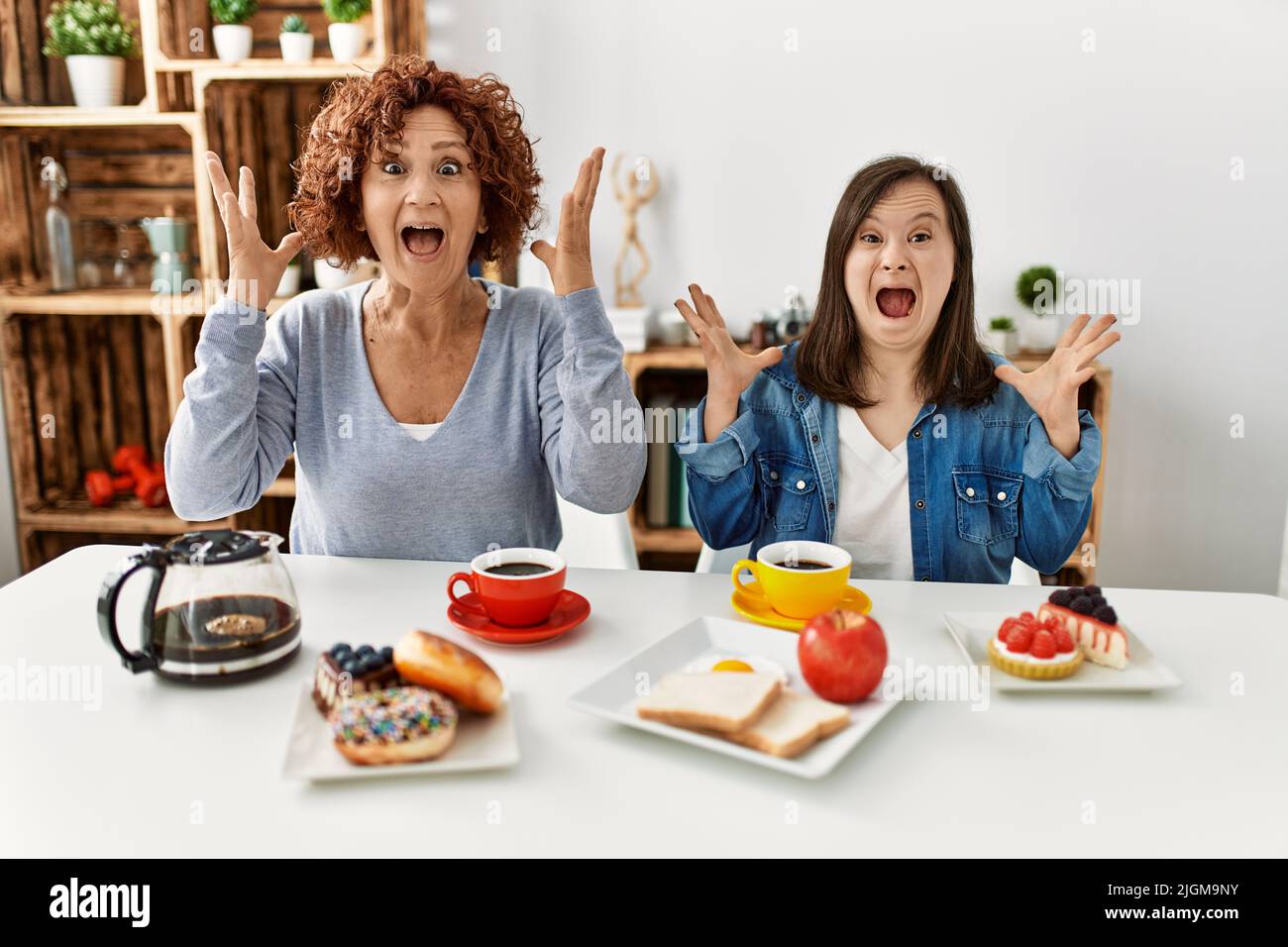 Family of mother and down syndrome daughter sitting at home eating breakfast celebrating crazy and amazed for success with arms raised and open eyes s Stock Photo