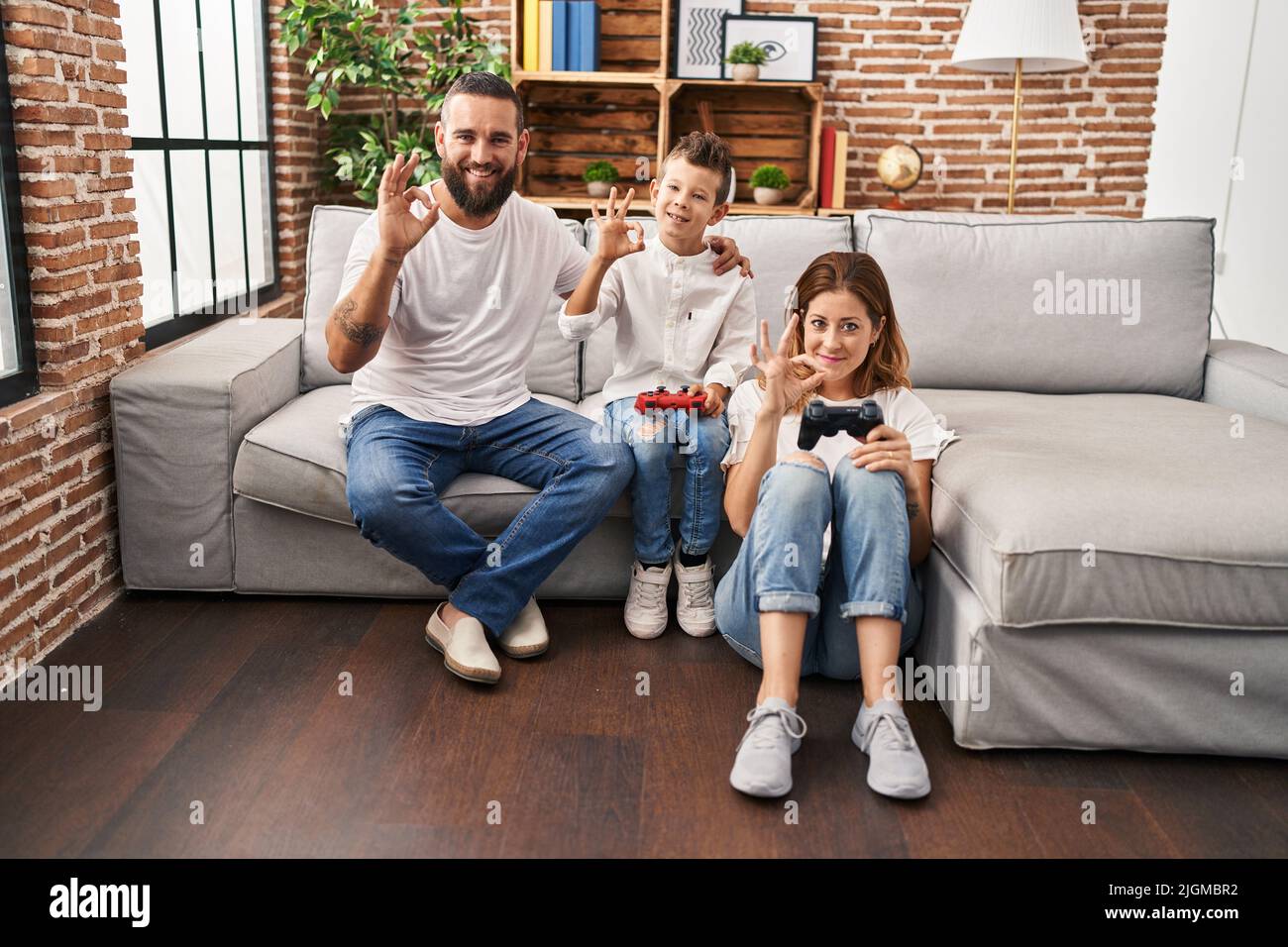 Family of three playing video game sitting on the sofa doing ok sign with fingers, smiling friendly gesturing excellent symbol Stock Photo