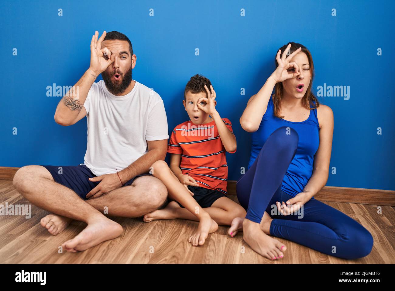 Family of three sitting on the floor at home doing ok gesture shocked with surprised face, eye looking through fingers. unbelieving expression. Stock Photo