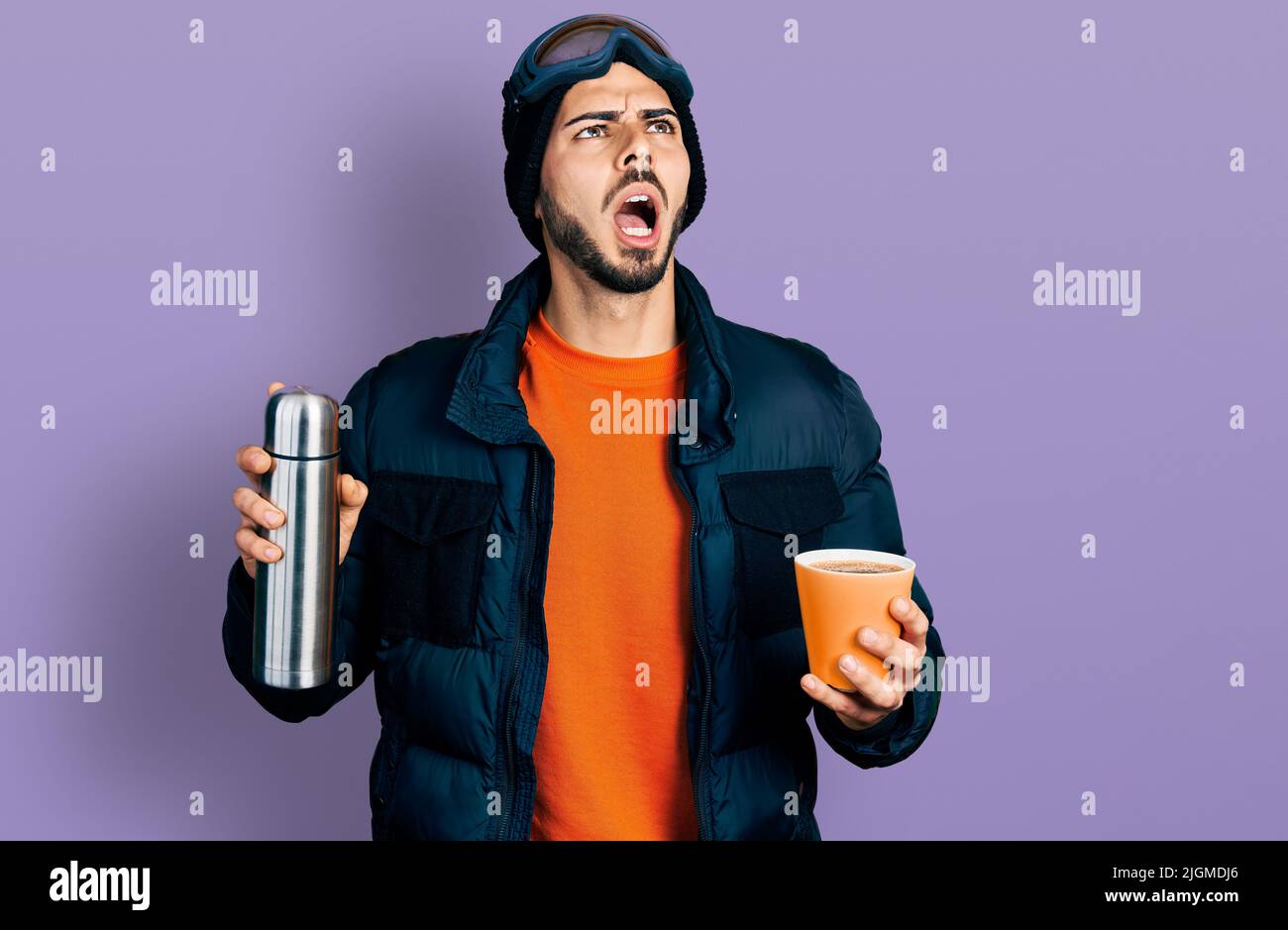 Young hispanic man with beard wearing snow wear and sky glasses holding coffee and thermo angry and mad screaming frustrated and furious, shouting wit Stock Photo