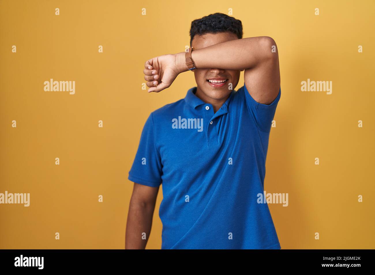 Young hispanic man standing over yellow background covering eyes with ...