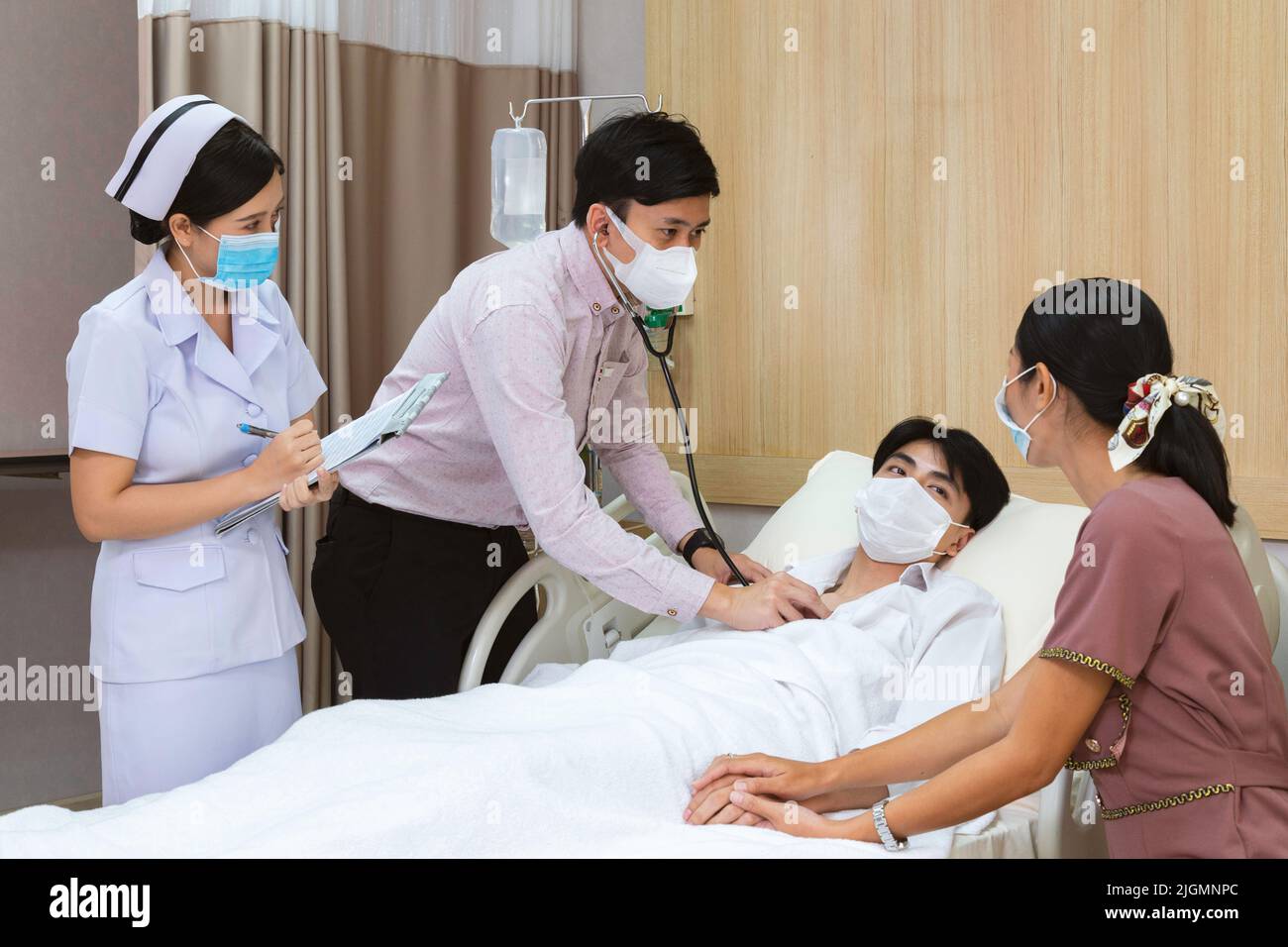 worrying asian mother holding hand to take care man patient while doctor check heartbeat by stethoscope and woman nurse writing health checklist at th Stock Photo