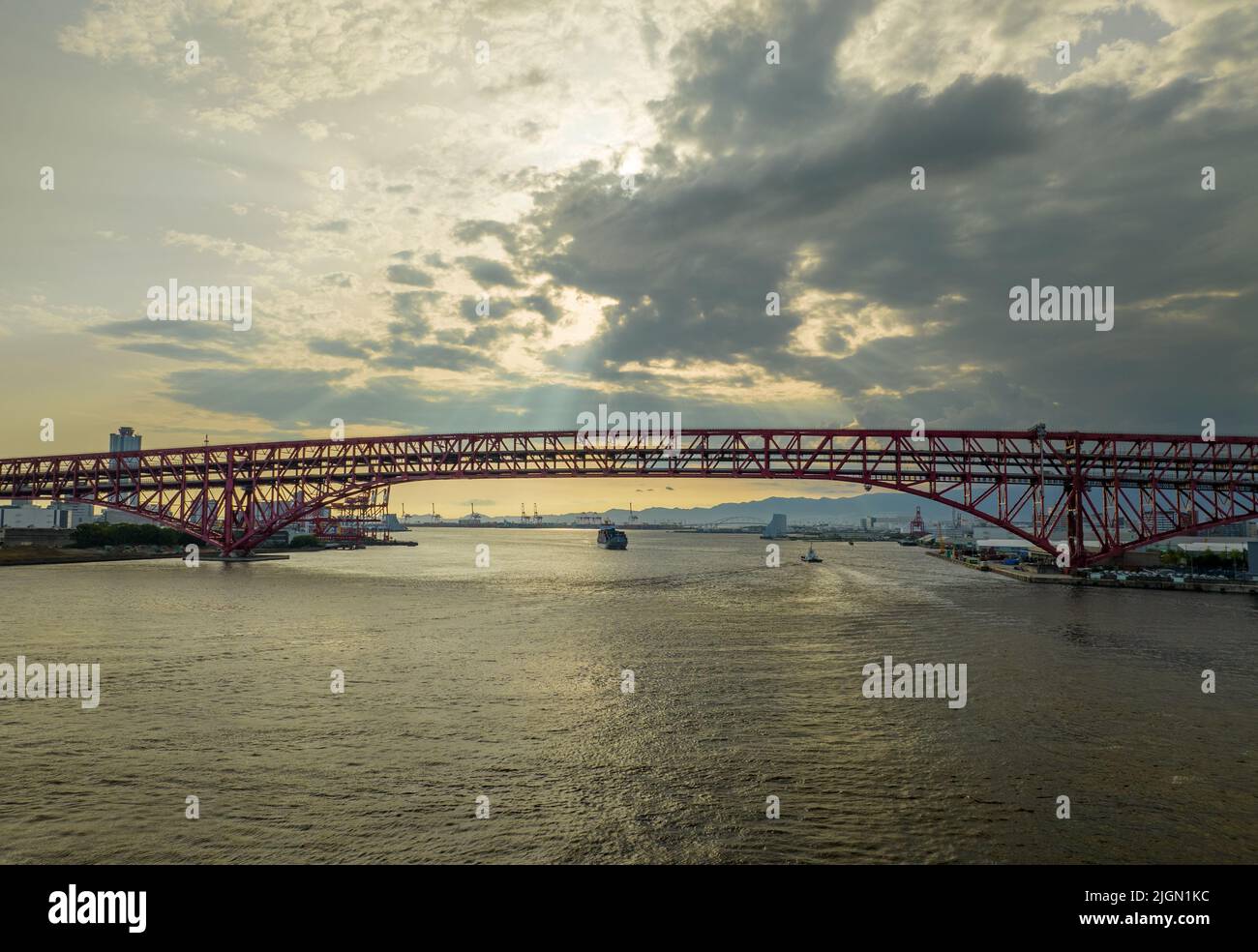 Sun shines through clouds on bridge over river and distant cargo ship Stock Photo