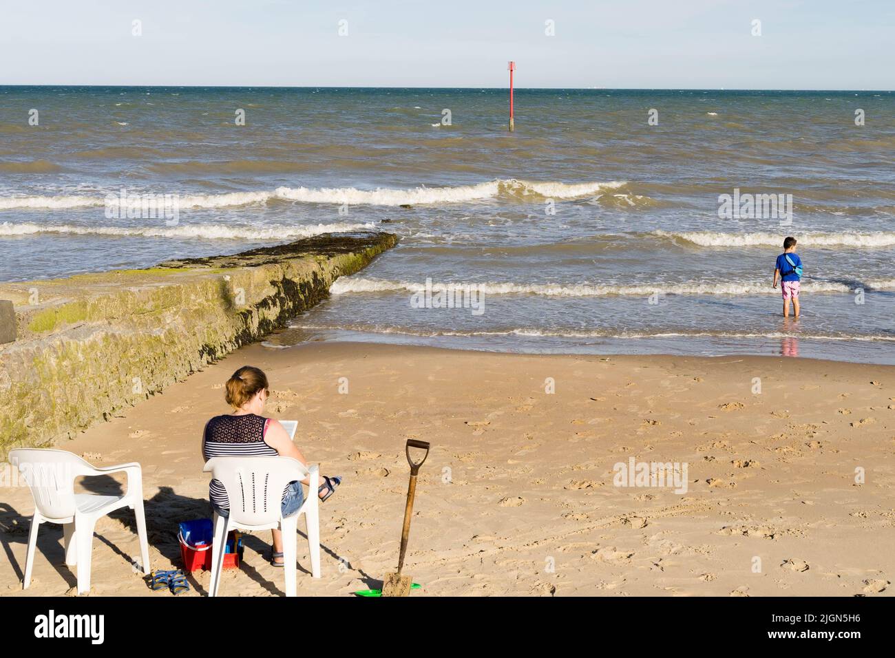 woman enjoys a quiet moment reading a book while her sun is playing with rising tidal wave on sandy beach in Kent England UK Stock Photo
