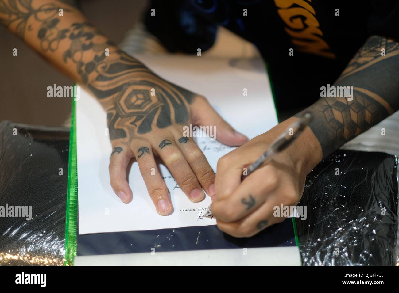 Overhead shot of tattooer, arms filled with graphic tattoos, as he stencils cursive script onto white paper with a pen in preparation for a session. Stock Photo