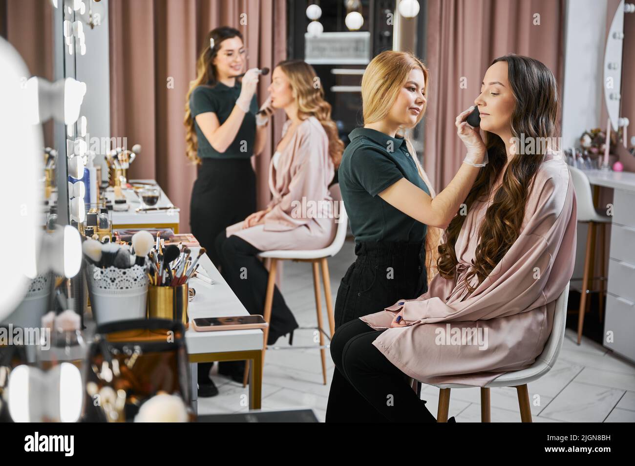 Makeup artists doing professional makeup for women in visage studio. Young woman beauty specialist in sterile gloves applying powder on client face with cosmetic brush. Stock Photo