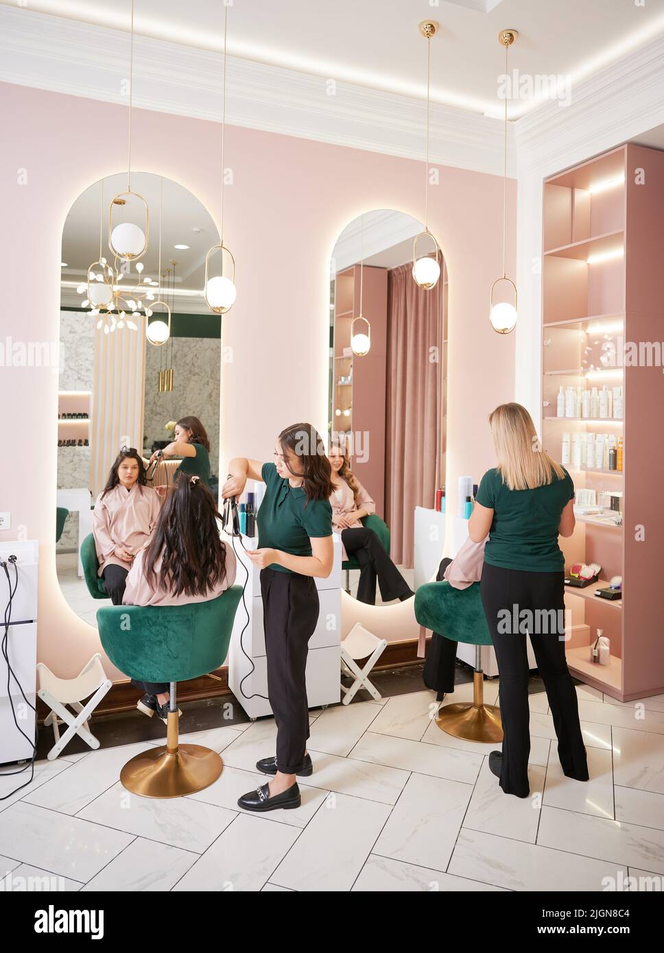 Two female hairstylists curling client hair and doing hairstyle in modern beauty salon. Young women sitting in chairs in front of mirrors while hairdressers styling clients hair. Stock Photo