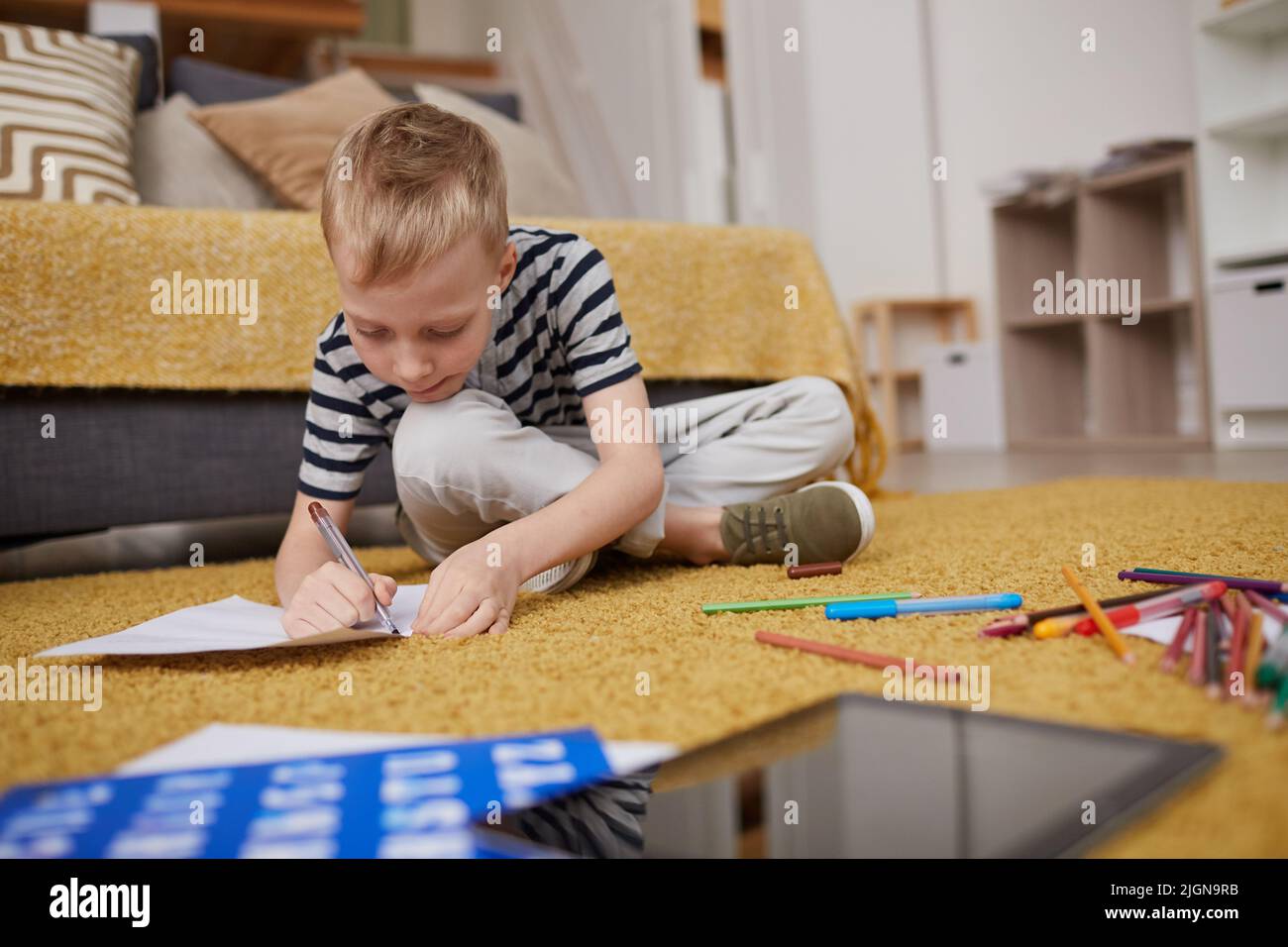 Busy blond-haired boy in casual outfit sitting with crossed legs on floor with pencils and drawing on paper in living room Stock Photo