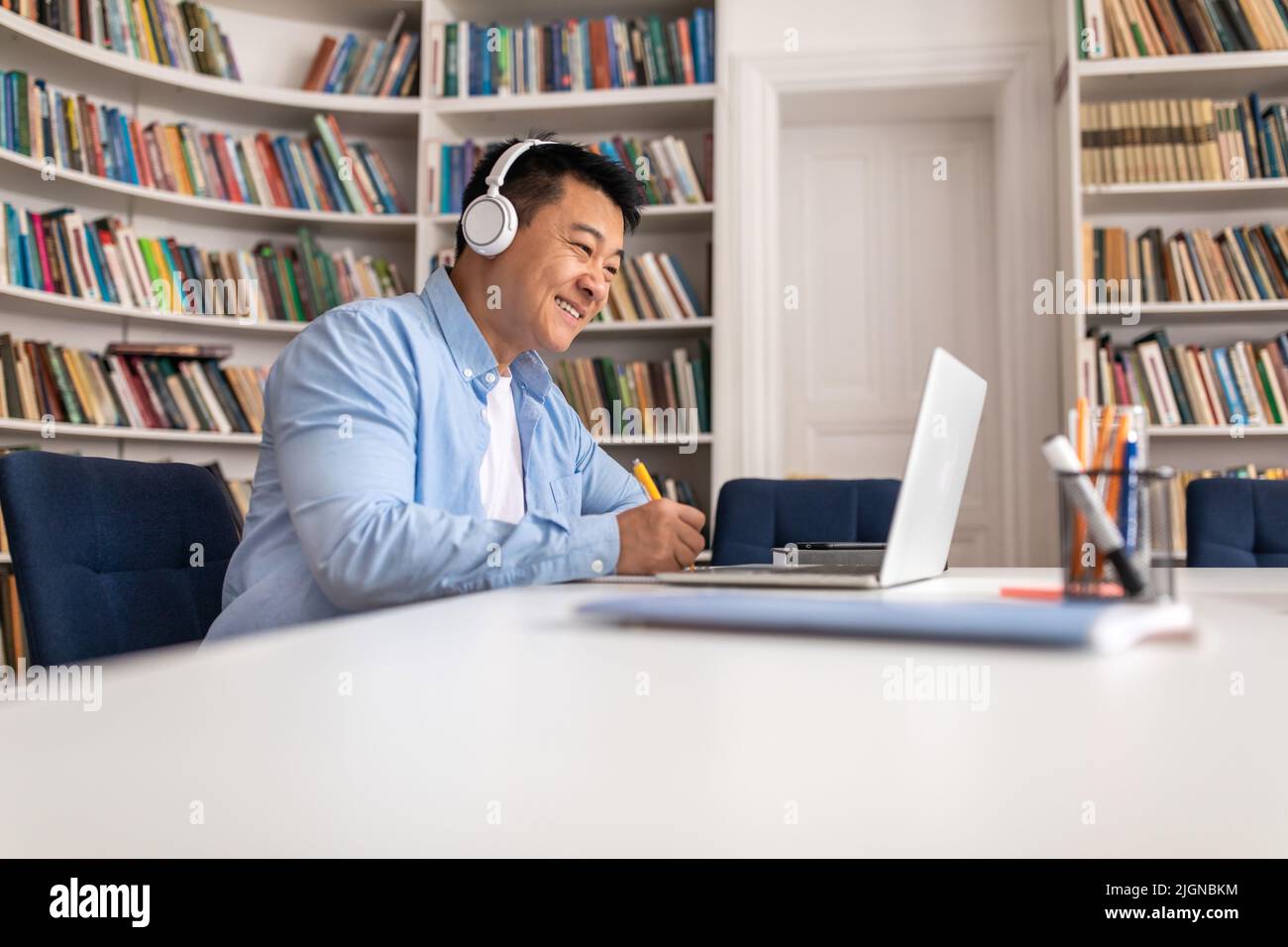 Happy Asian Man Using Laptop And Taking Notes In Library Stock Photo