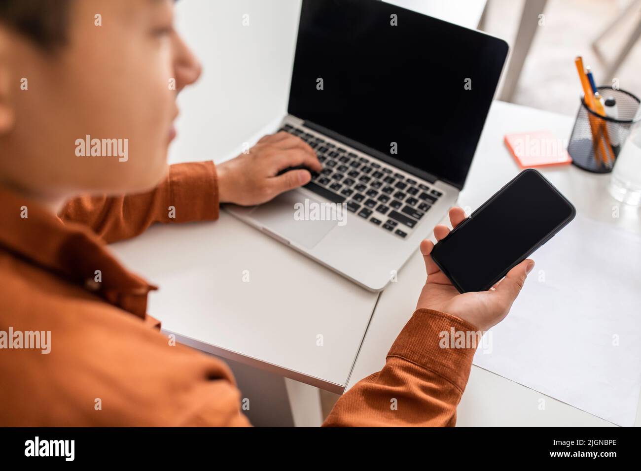 Asian Businessman Using Smartphone And Laptop With Empty Screens Indoor Stock Photo