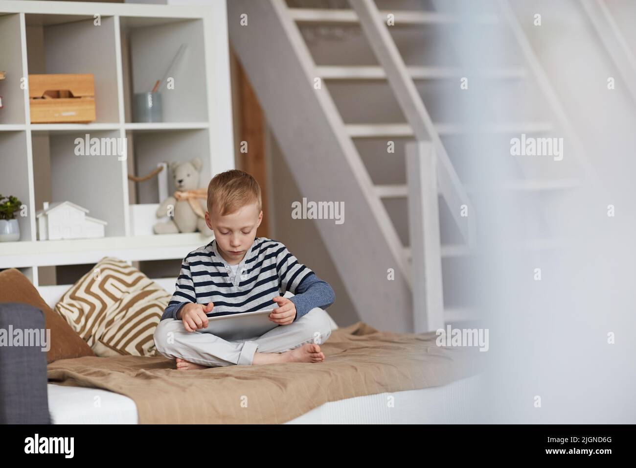 Serious little boy with blond hair sitting with crossed legs on bed and