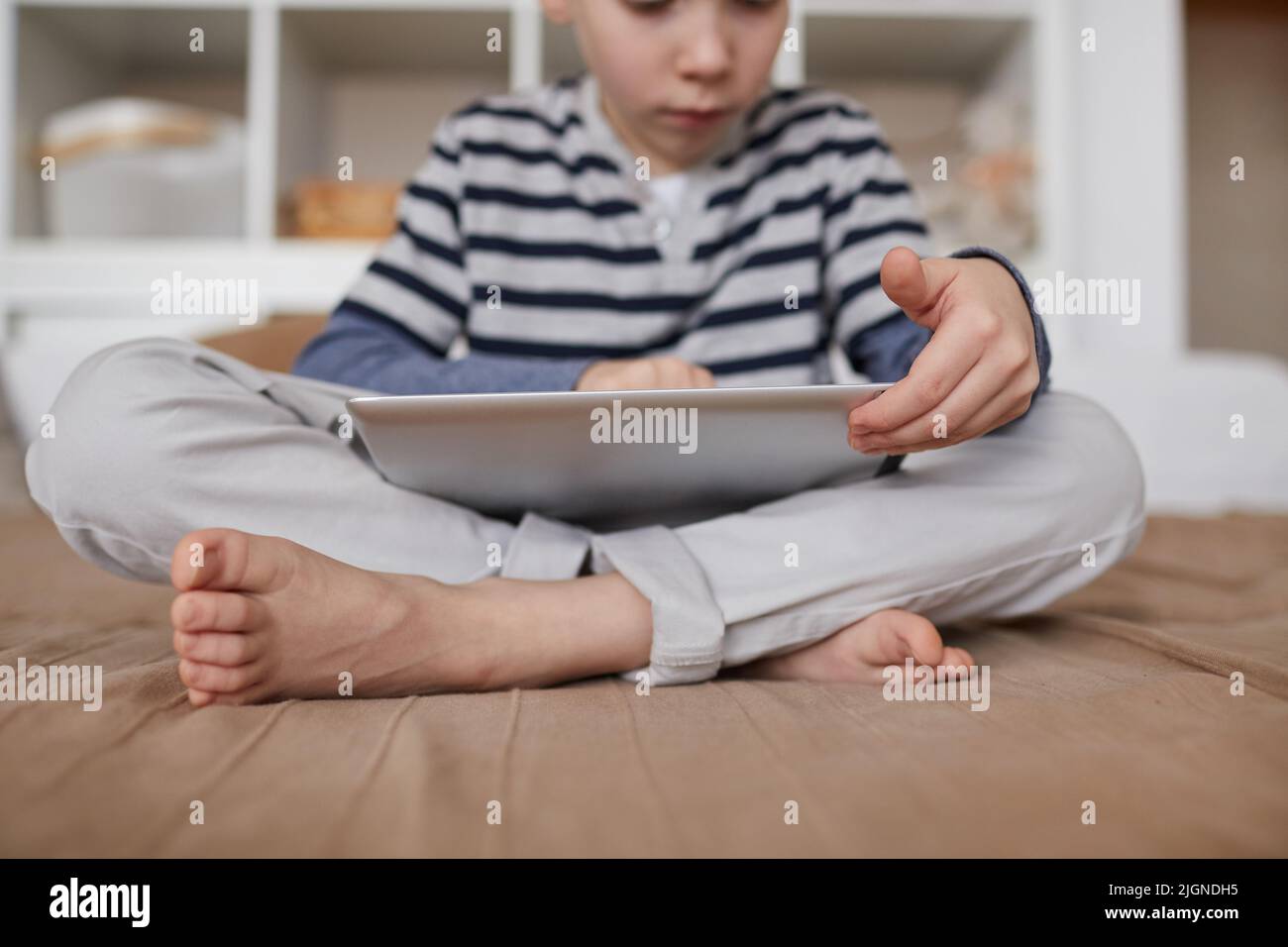 Close-up of curious barefoot child sitting with crossed legs on bed and scrolling on tablet Stock Photo
