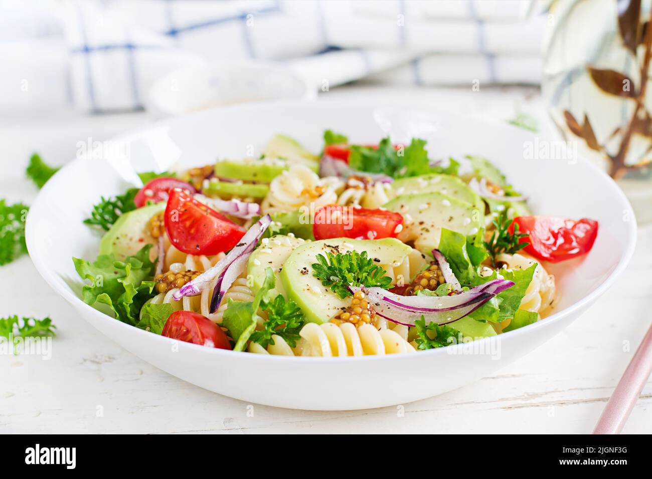 Fusilli pasta salad with avocado, tomatoes, fresh green lettuce, red onion and mustard dressing on white background. Vegetarian healthy lunch. Stock Photo