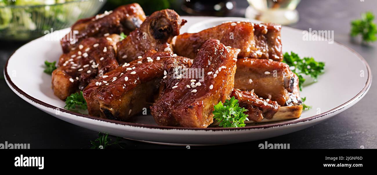 Delicious barbecued spare ribs on plate on dark background. Tasty bbq meat. Banner Stock Photo