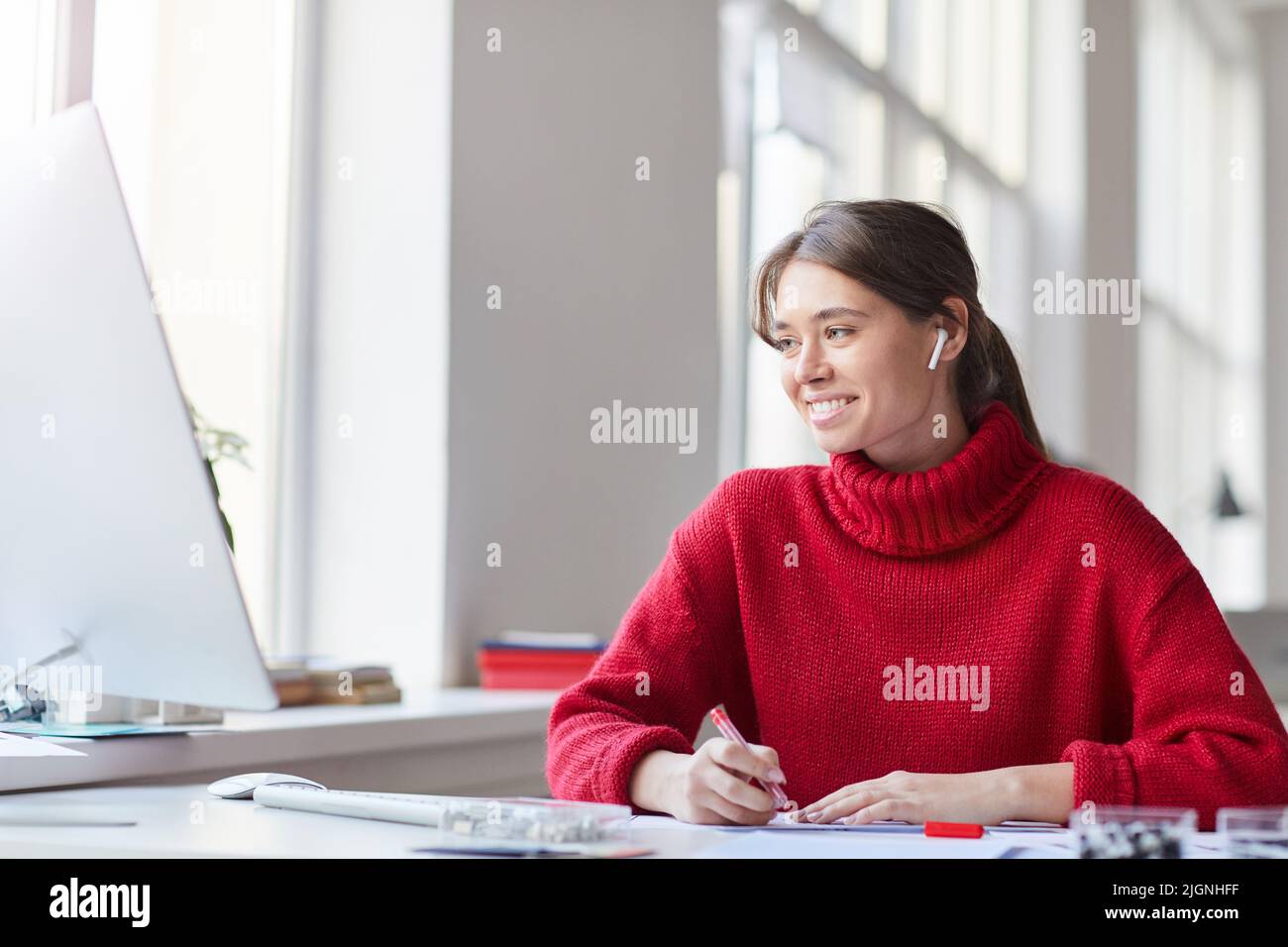 Smiling attractive young woman in red knitted sweater and earbuds sitting at table and using computer while working on web design project Stock Photo