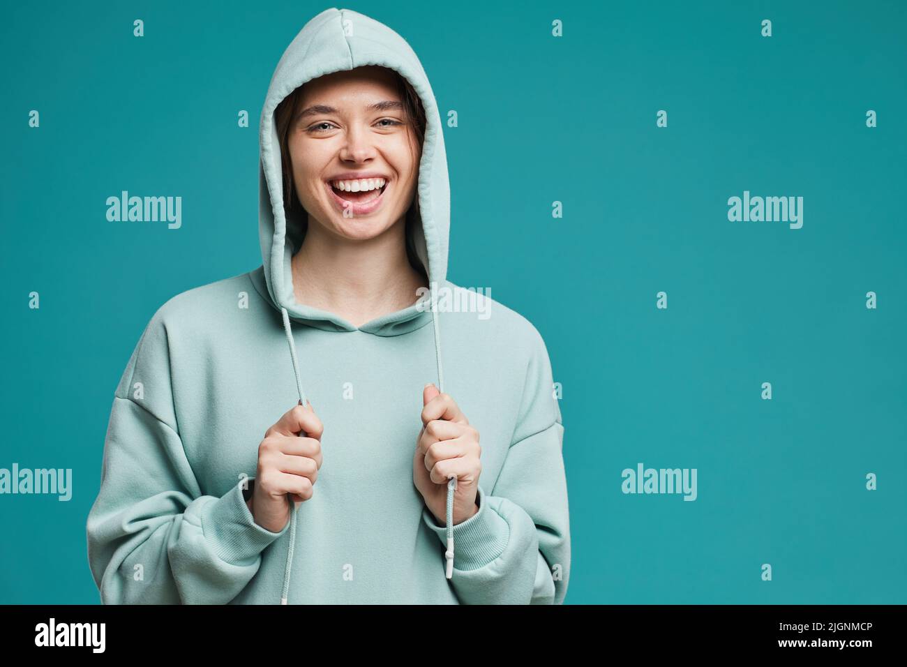 Portrait of happy beautiful girl in teal hoodie pulling strings down and laughing cheerfully against blue background Stock Photo