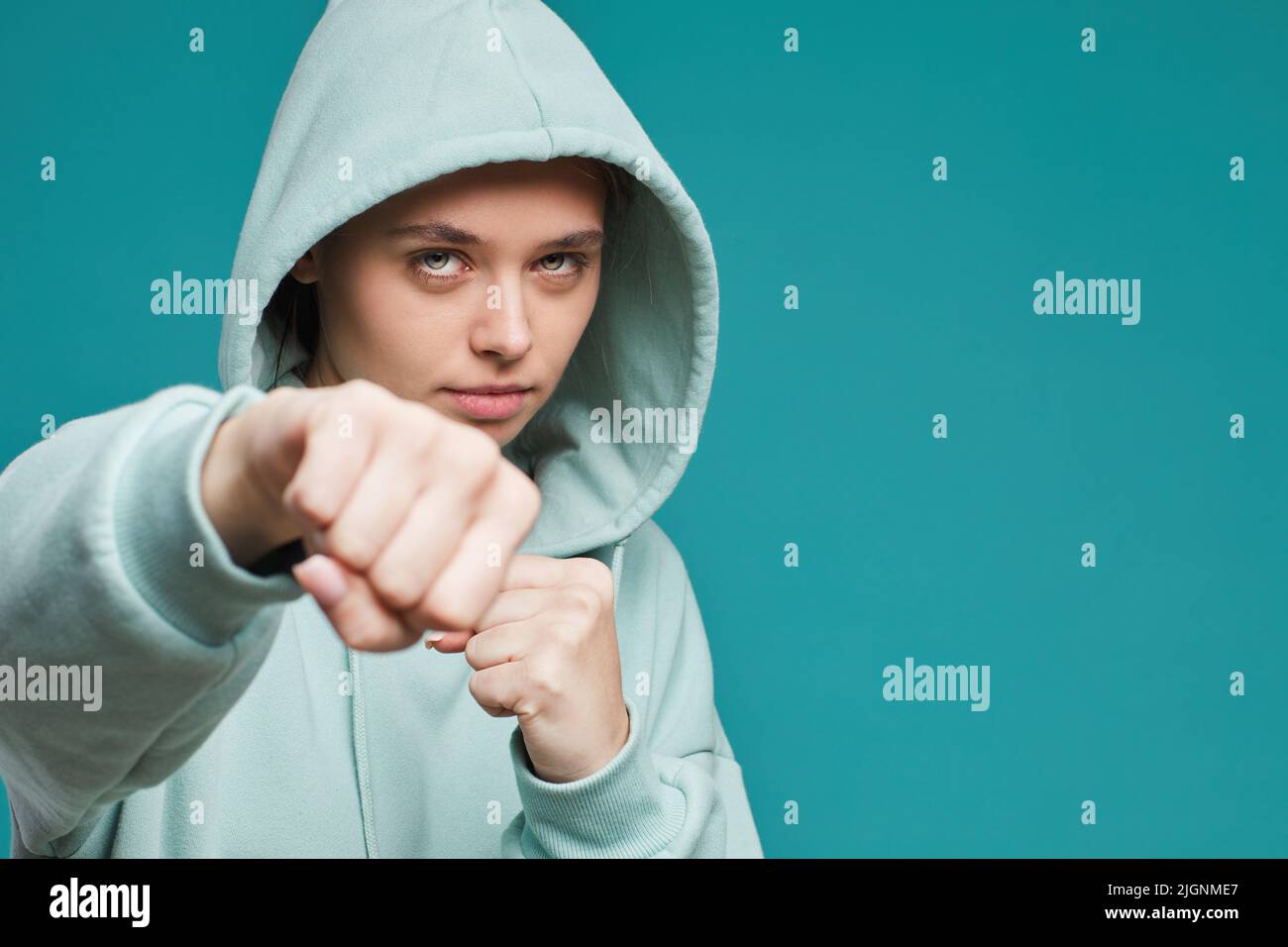 Portrait of serious female fighter in teal hoodie punching at camera while preparing for fight, isolated background Stock Photo