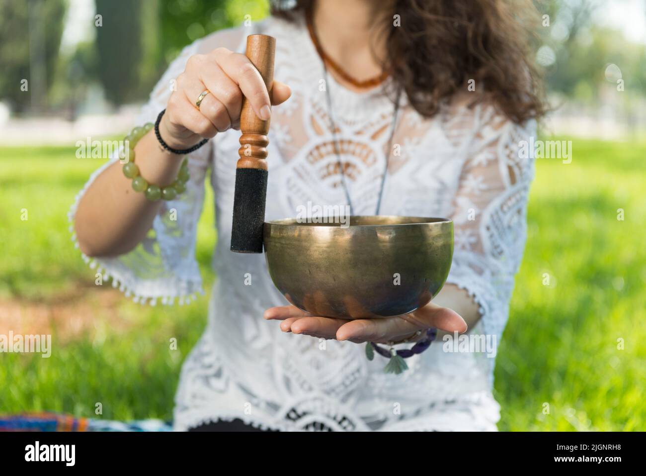 Detail of a woman's hands holding and playing a Tibetan singing bowl during a meditation and music therapy session in a park area. Stock Photo