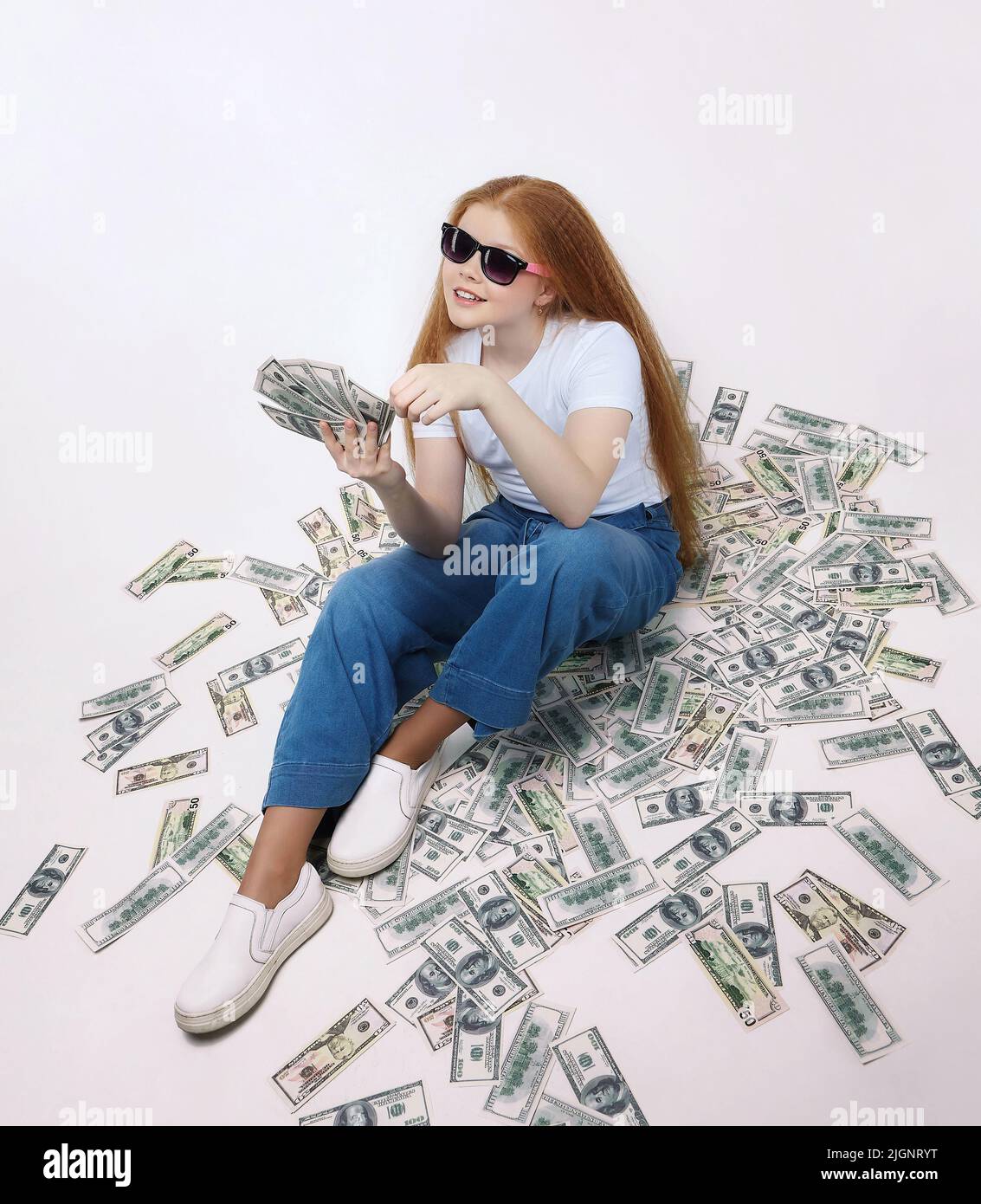 beautiful red-haired girl is sitting on money. Dollar bills are scattered on the floor. Stock Photo