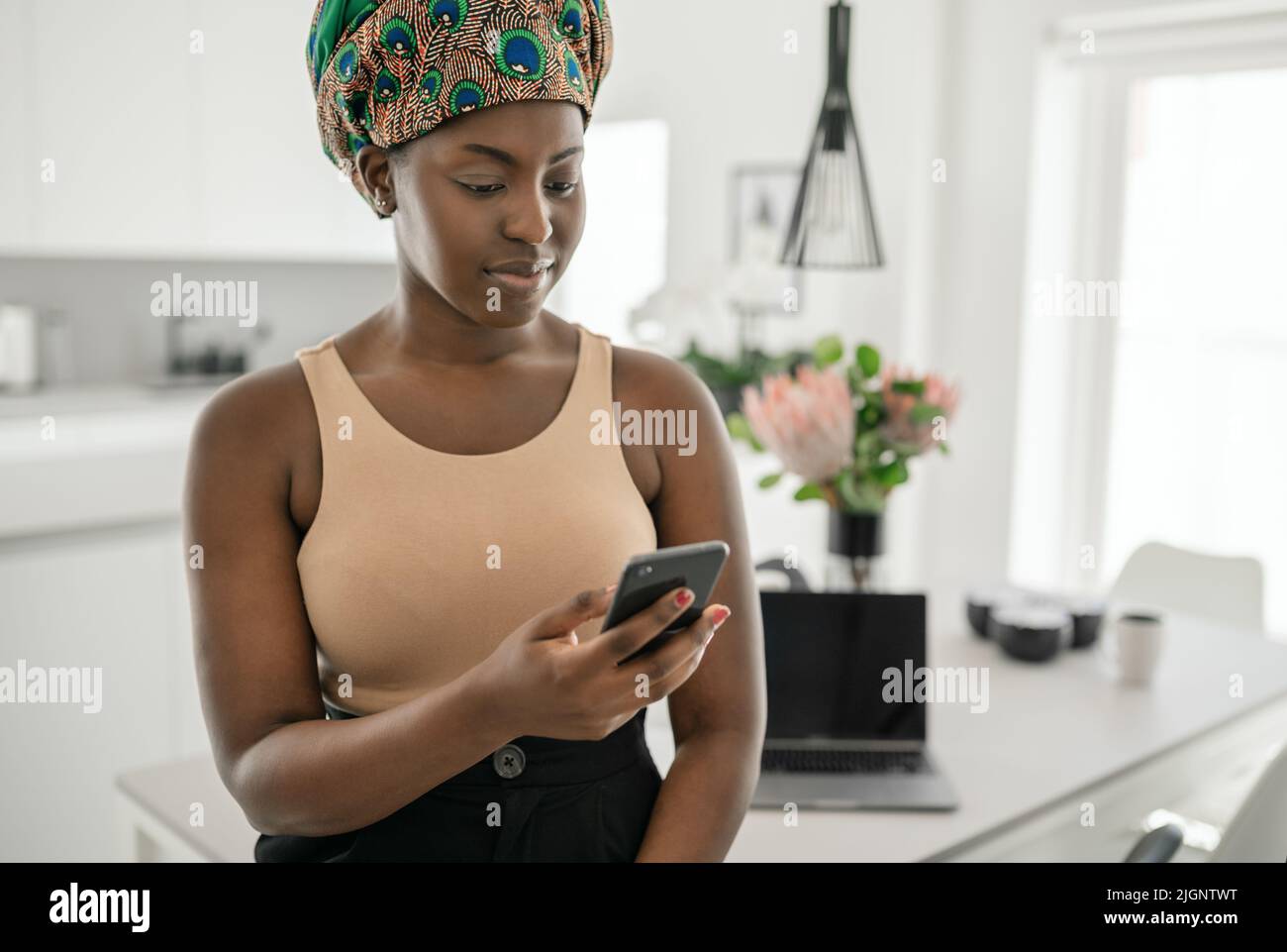 Beautiful Black African woman working at home, smiling, using mobile smart phone, wearing traditional headscarf Stock Photo