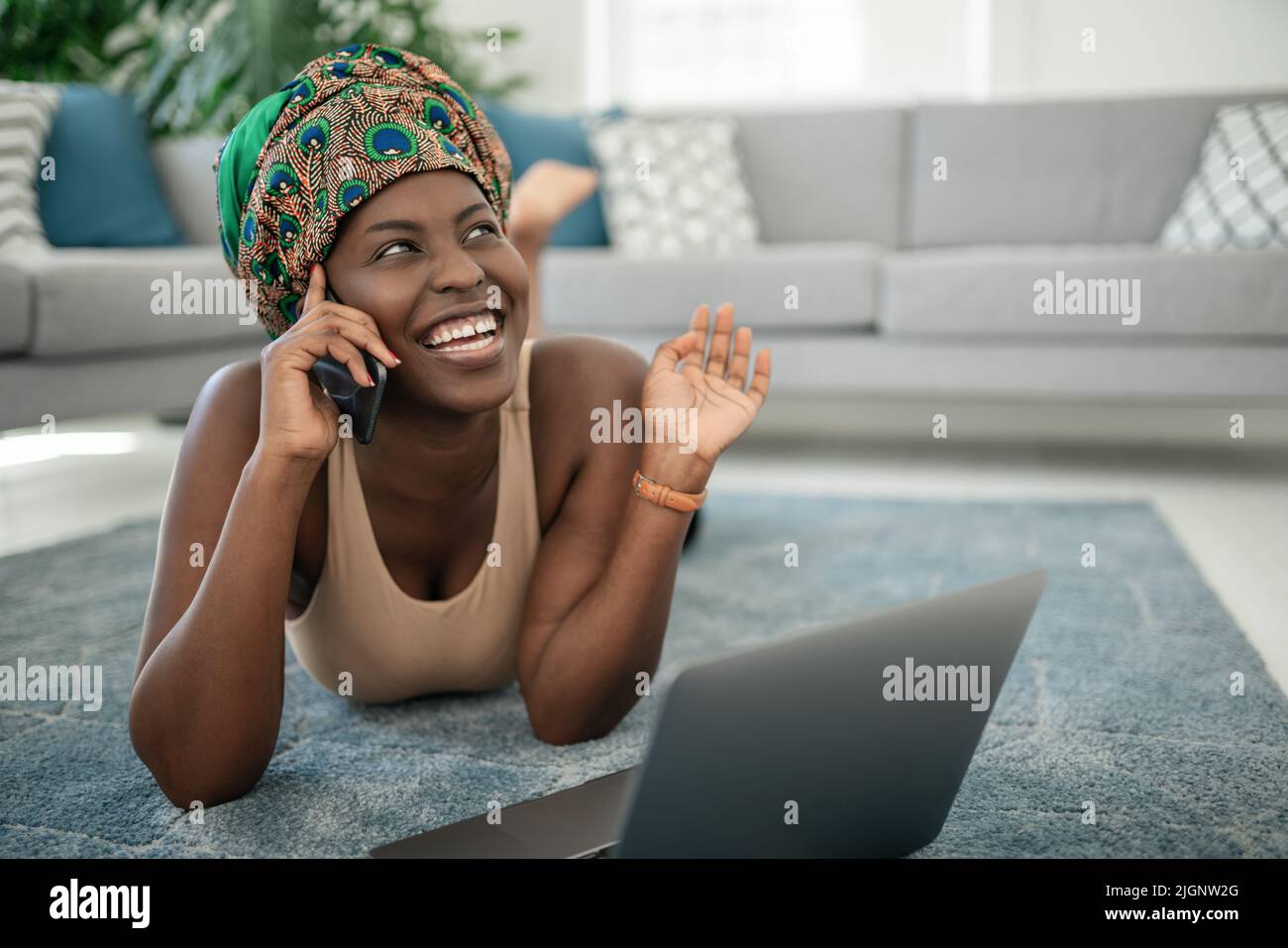 Beautiful African woman at home, smiling and laughing, using mobile smart phone, wearing traditional headscarf, surprised and happy Stock Photo