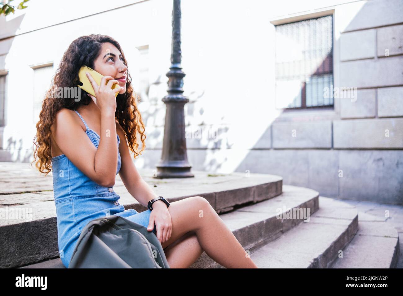 Young indian female tourist wearing denim dress talking by phone. She is sitting on steps at a Barcelona old town Stock Photo