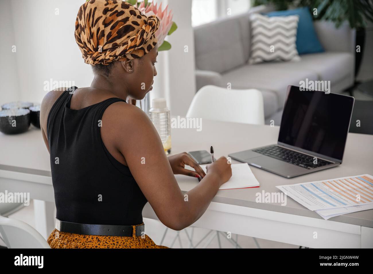 African woman studying from home, looking at laptop, writing down notes ...