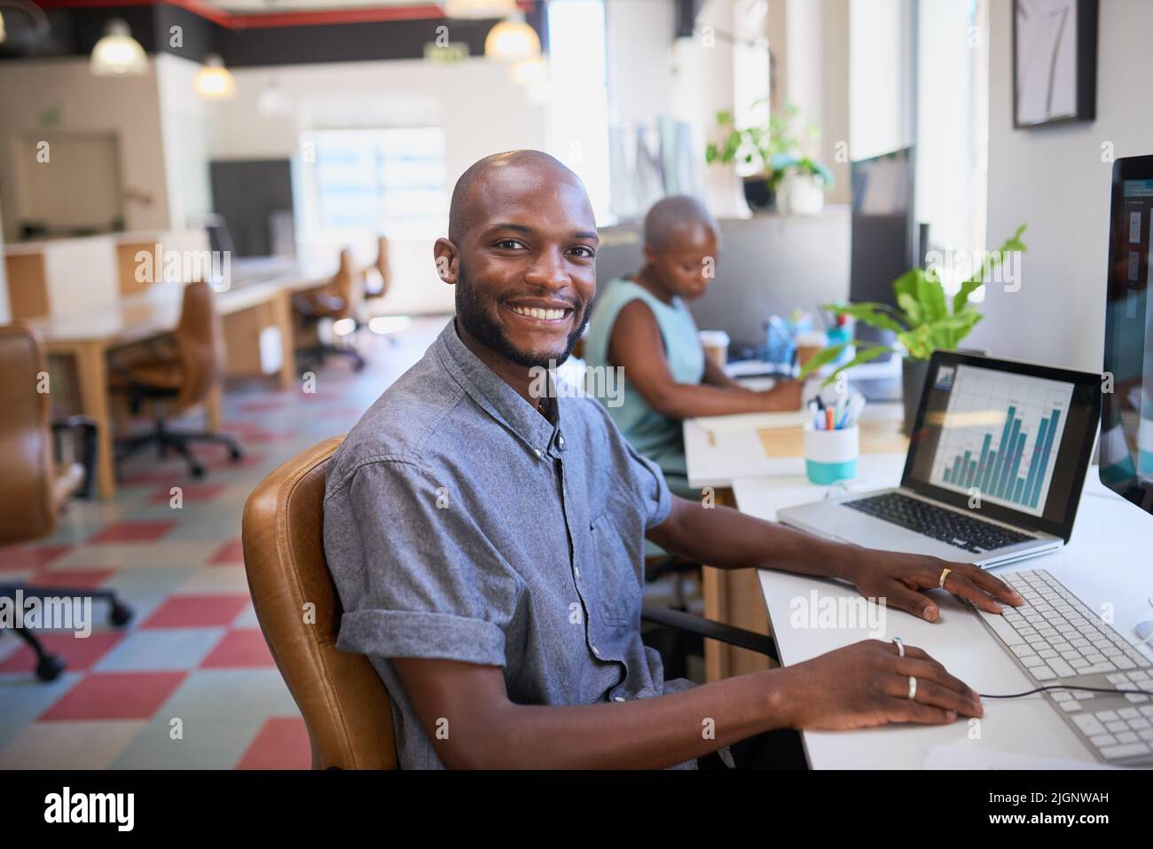 A Black man smiles at his desk in open plan creative office Stock Photo