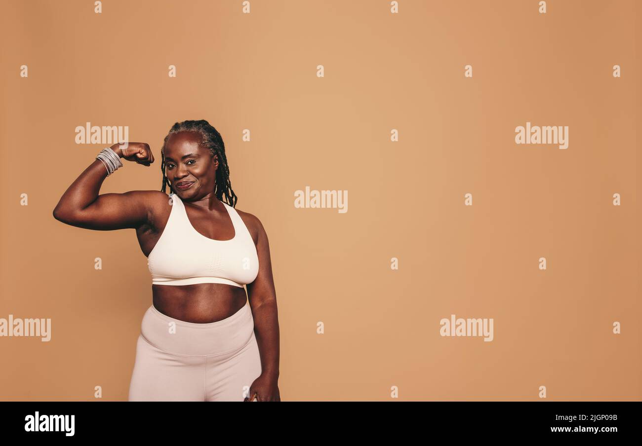 Woman with dreadlocks looking at the camera while flexing her bicep. Mature woman standing against a brown background in sports clothing. Sporty black Stock Photo