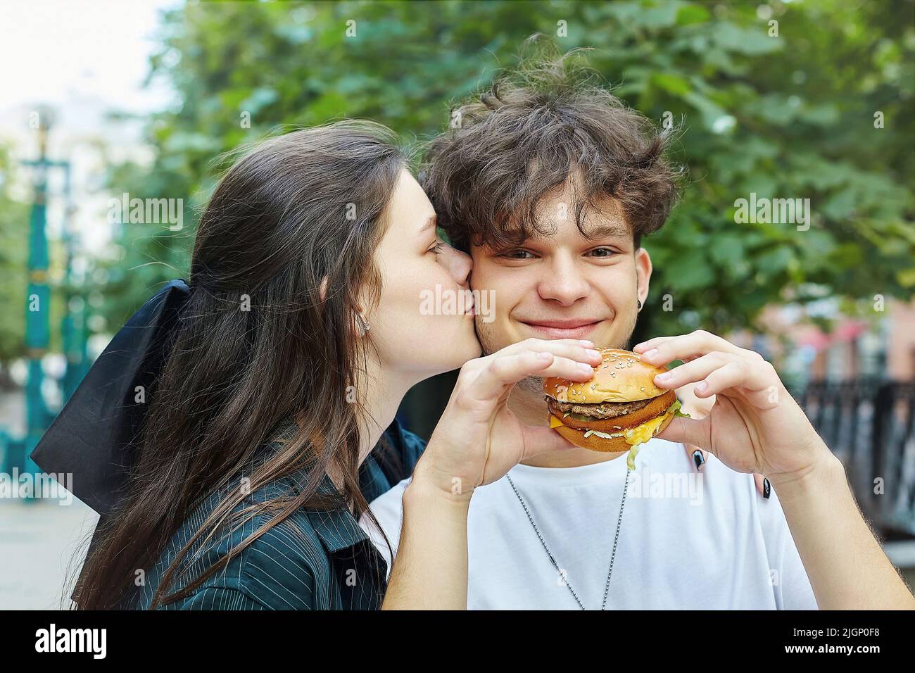 young girl kisses a guy who is holding a burger in his hands. Stock Photo
