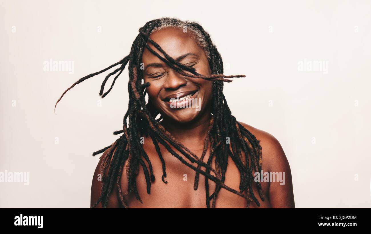 Cheerful woman smiling and whipping her dreadlocks while standing against a studio background. Mature black woman wearing a bath towel and make-up. Ha Stock Photo