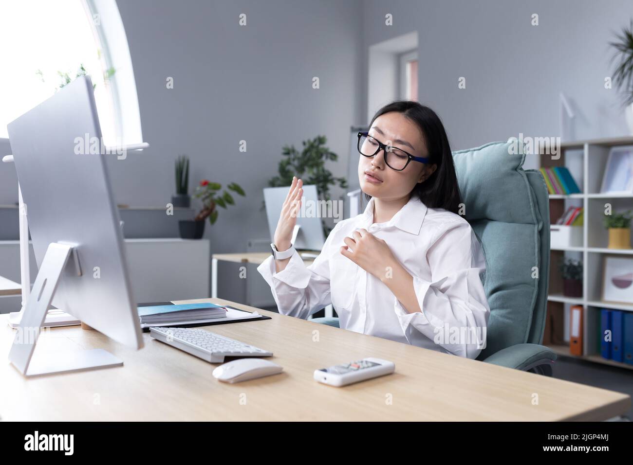 Heat in the workplace. Young Asian woman sitting in the office at the desk waving her hand, it's very hot, she feels bad, she needs fresh air, take a break. Stock Photo