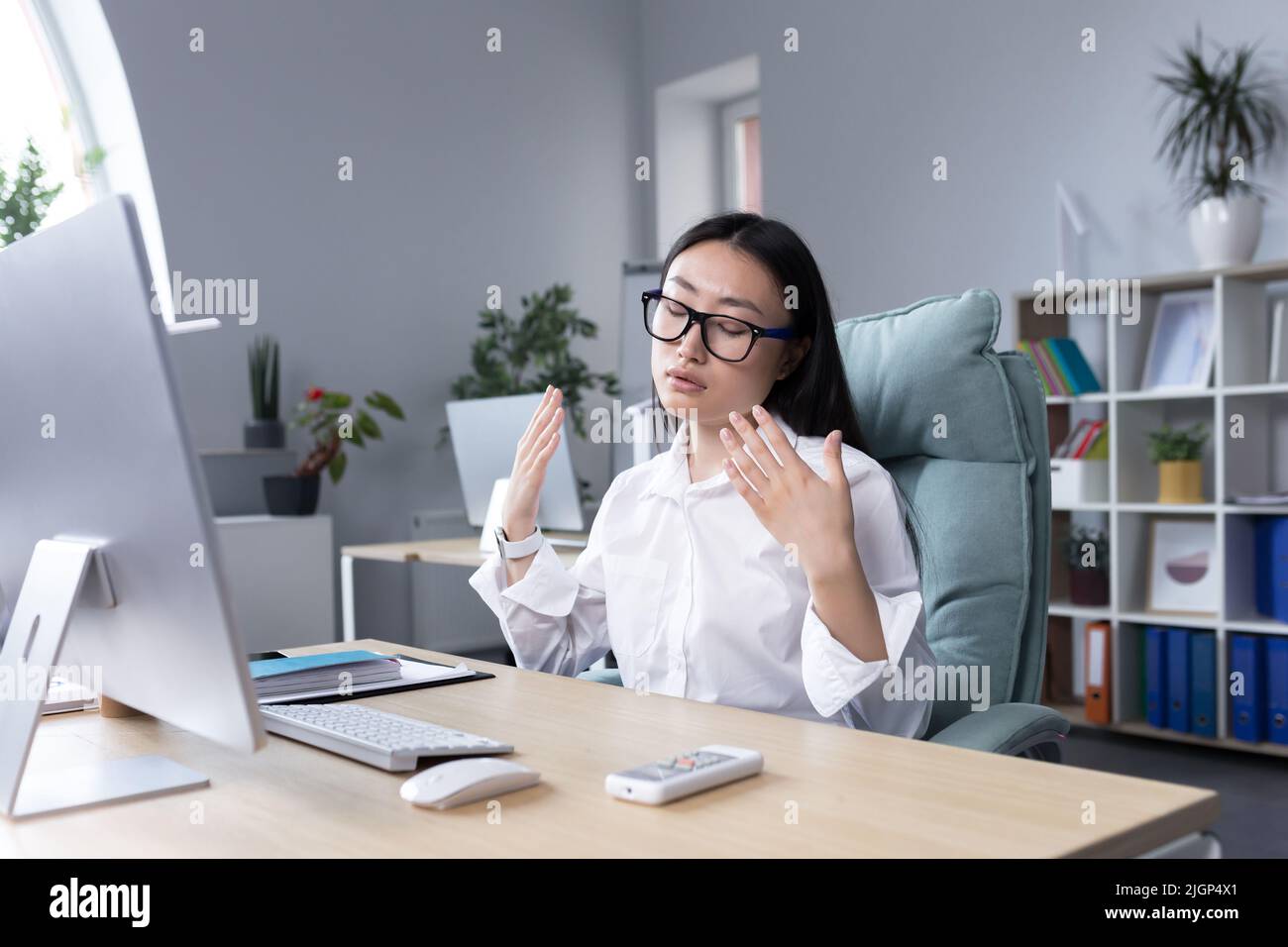 Heat in the workplace. A young Asian woman sits in the office at the desk, waves both hands at her face, it is very hot, she feels bad, she needs fresh air, take a break. Stock Photo