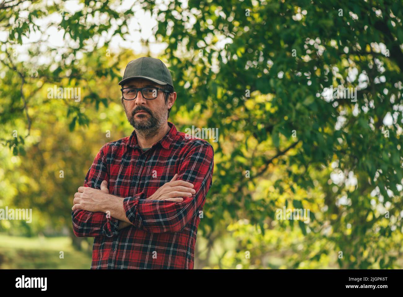 Portrait of male farmer with arms crossed wearing plaid shirt and trucker's hat posing in walnut orchard and looking at camera, selective focus Stock Photo