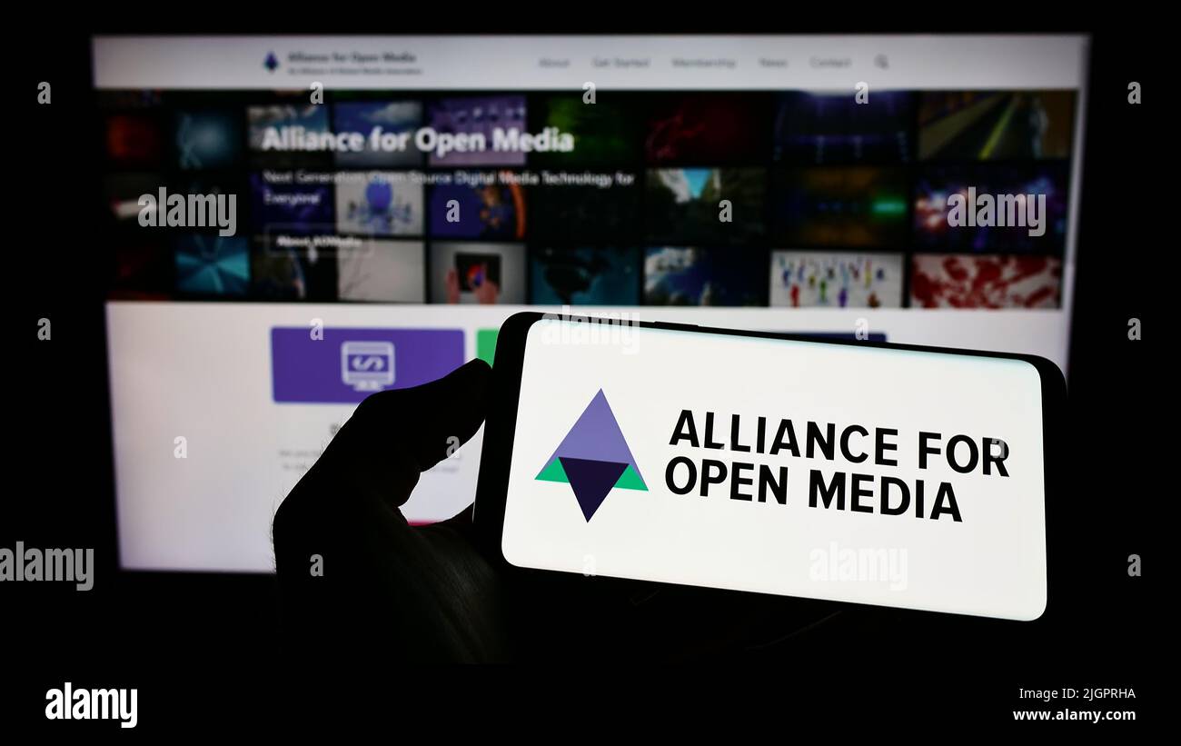 Person holding cellphone with logo of consortium Alliance for Open Media (AOMedia) on screen in front of business webpage. Focus on phone display. Stock Photo