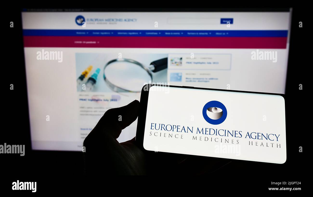 Person holding mobile phone with logo of EU agency European Medicines Agency (EMA) on screen in front of web page. Focus on phone display. Stock Photo